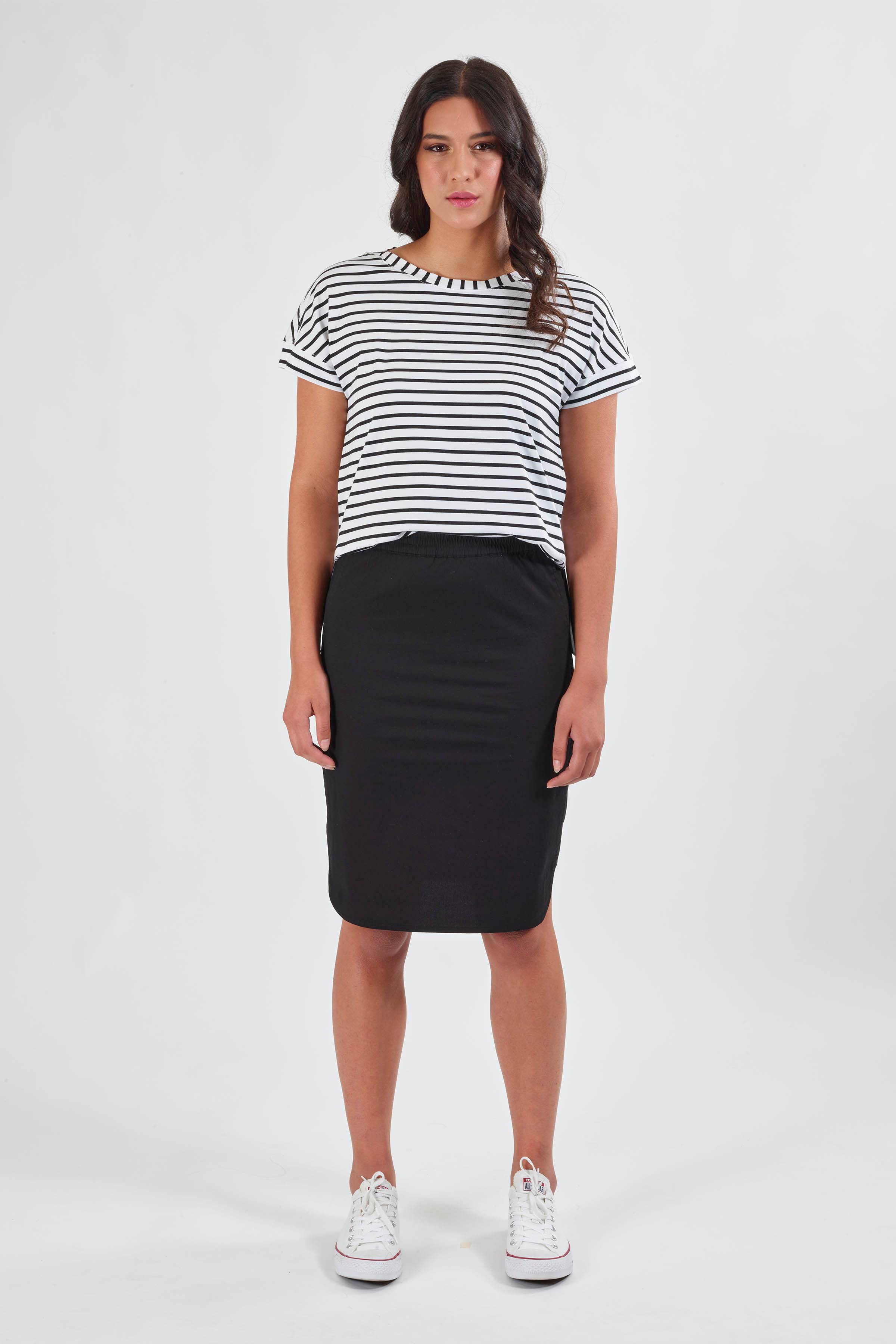 A woman faces forward against a plain white background, wearing the Vassalli Poplin Comfort Skirt in black, a black-and-white striped short-sleeve shirt, and white sneakers.