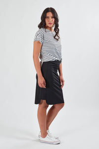 A woman with long wavy hair and a neutral expression models the Vassalli Poplin Comfort Skirt in black, paired with a white and black striped short-sleeve top and white sneakers against a plain white background.