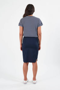 A woman stands facing away against a white background, wearing a navy blue and white striped short-sleeve top, Vassalli’s Poplin Comfort Skirt - Navy, and white sneakers.