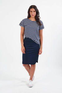 A woman with long dark hair and a relaxed expression wears the Vassalli Poplin Comfort Skirt in navy, paired with a navy and white striped t-shirt and white sneakers, standing against a white background.