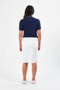A woman, facing away from the camera, wears a navy short-sleeve polo, Vassalli’s Poplin Comfort Skirt in white, and white slip-on shoes against a plain white background.