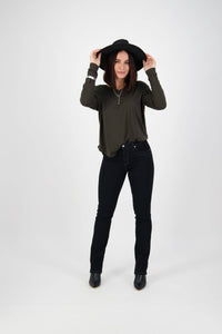 A woman in a black hat, dark long-sleeve shirt, Vassalli Studio Jean - Black Denim, and black boots stands smiling against a white background, holding her hat brim with both hands.
