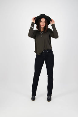 A woman in a black hat, dark long-sleeve shirt, Vassalli Studio Jean - Black Denim, and black boots stands smiling against a white background, holding her hat brim with both hands.
