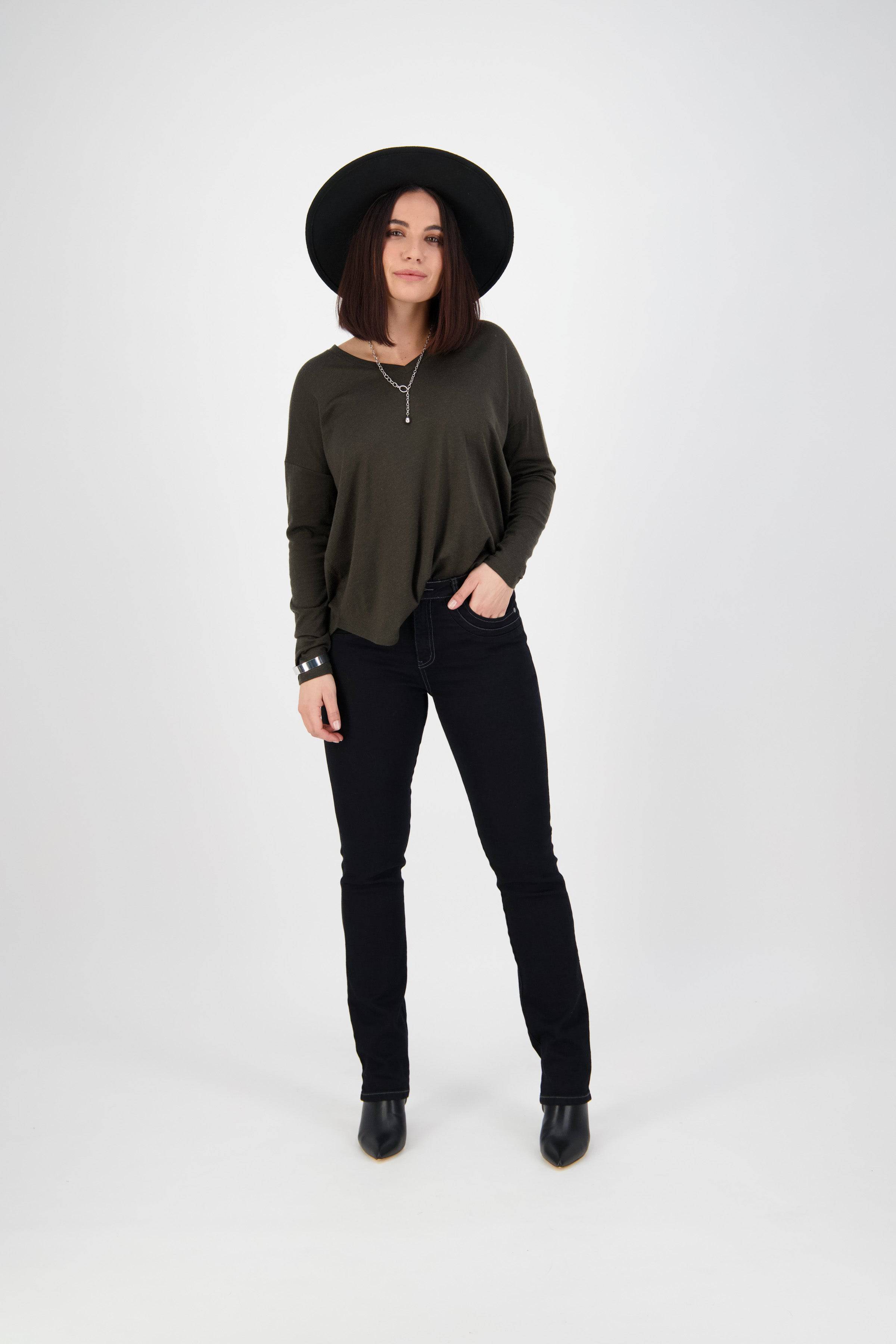 A woman stands against a plain white background, wearing a black wide-brimmed hat, an olive green long-sleeve shirt, Vassalli Studio Jean in Black Denim, and black ankle boots. She has one hand in her pocket and looks relaxed.