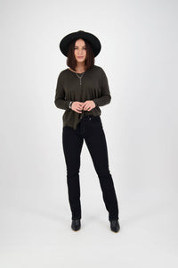 Woman wearing a wide-brimmed black hat, dark green long-sleeve top, Vassalli Studio Jean - Black Denim, and black ankle boots stands against a plain white background.