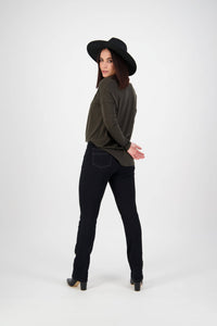 A woman in a wide-brimmed black hat, dark green top, and Vassalli Studio Jean - Black Denim stands with her back turned, looking over her shoulder with hands behind her back against a plain white background.