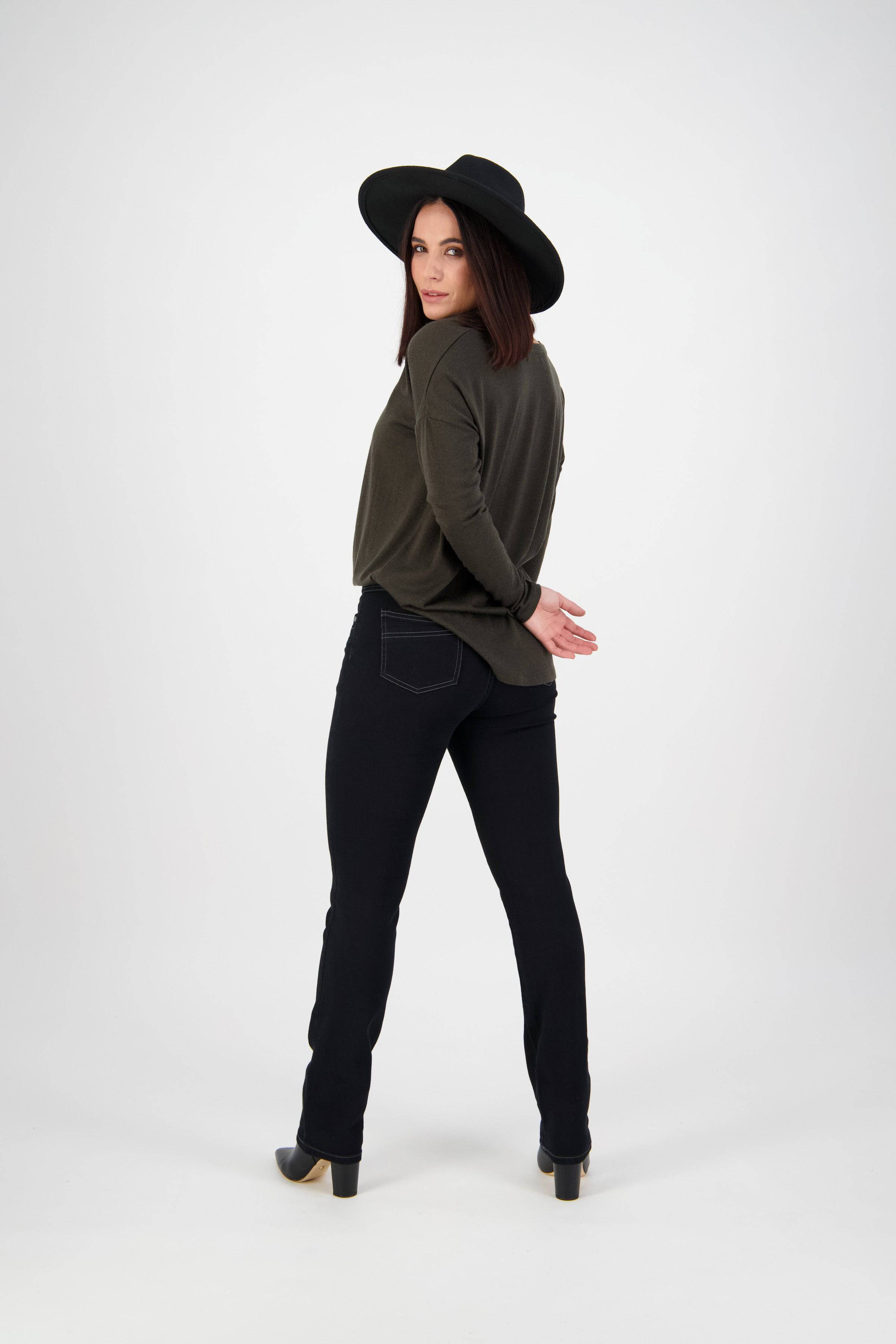 A woman in a wide-brimmed black hat, dark green top, and Vassalli Studio Jean - Black Denim stands with her back turned, looking over her shoulder with hands behind her back against a plain white background.