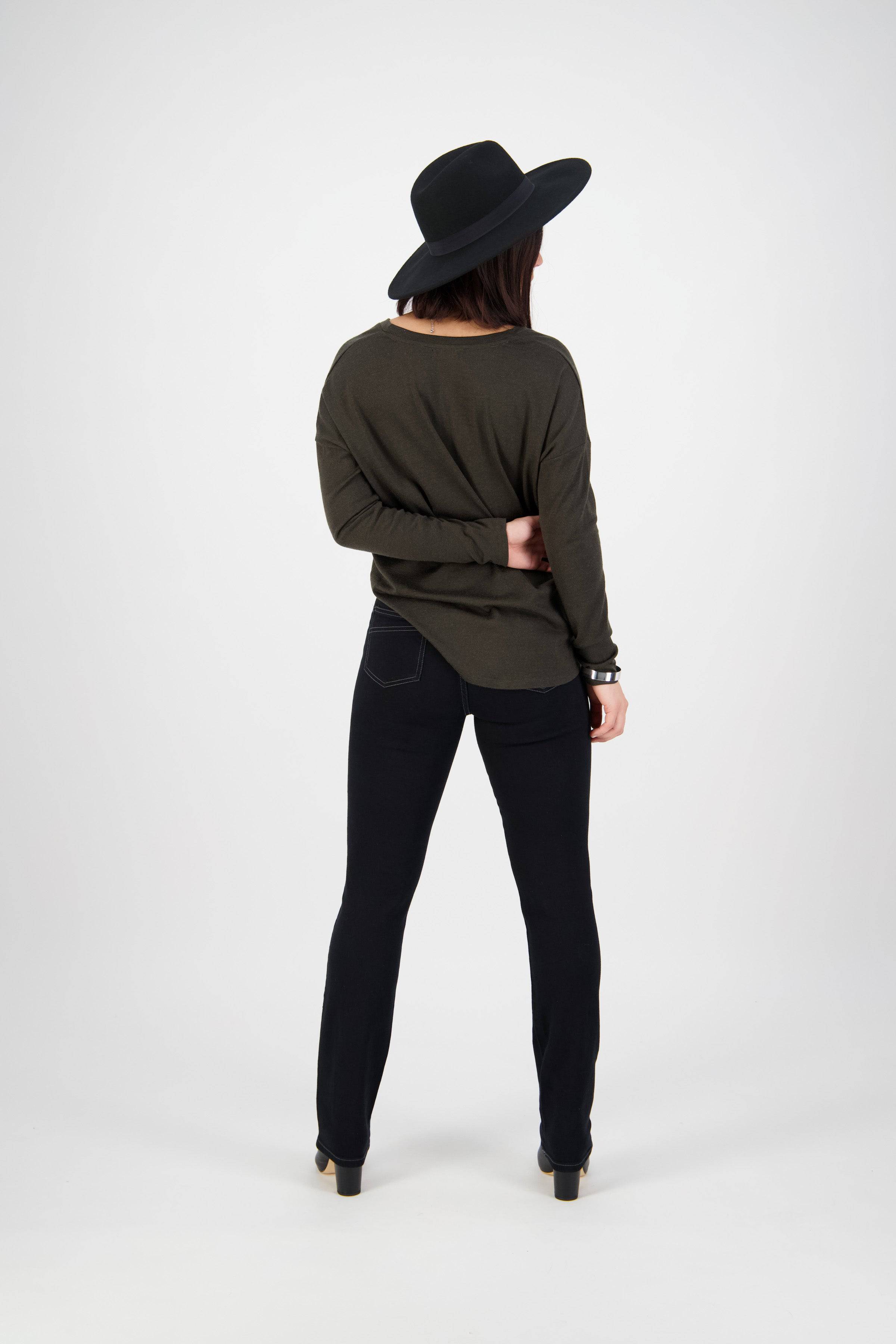 A person in a wide-brimmed black hat, long-sleeved olive shirt, and Vassalli Studio Jean - Black Denim stands facing away from the camera with one hand behind their back against a plain white background.