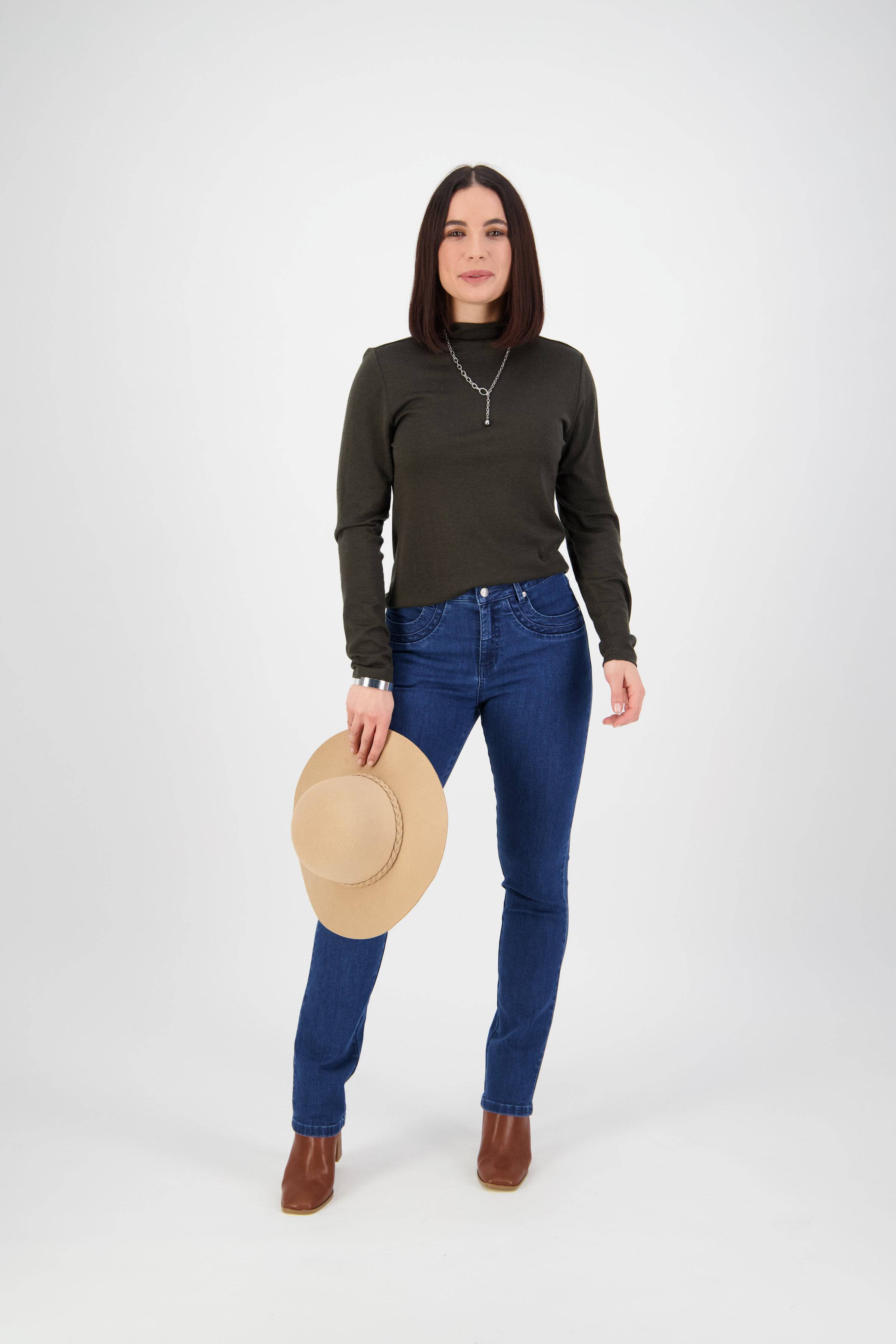 A woman with straight dark hair wears a dark long-sleeve top, Vassalli Studio Jean - Blue Denim, and brown boots. She stands on a white background, holding a beige hat in her left hand with a neutral expression.