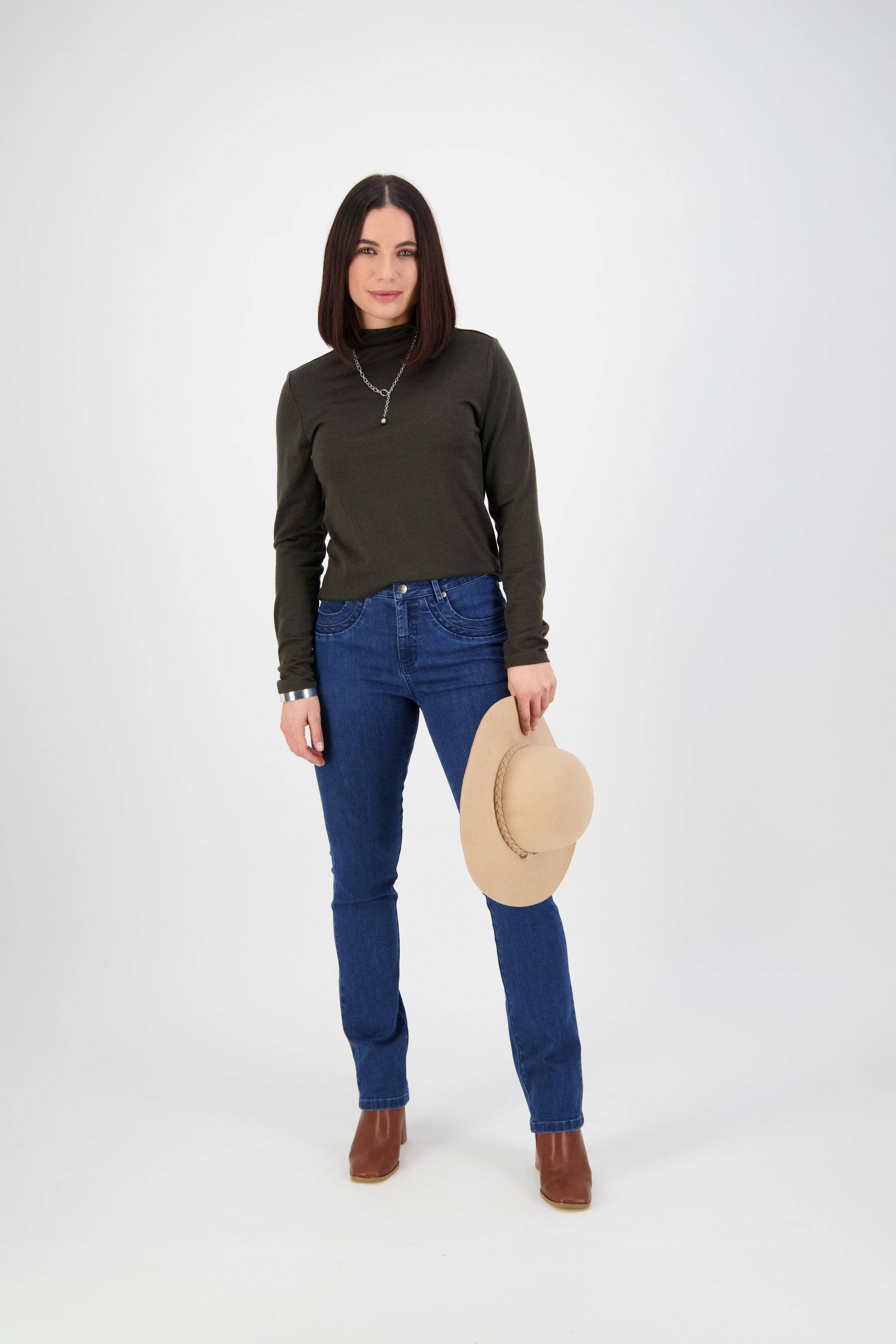 A woman with straight dark hair faces forward, wearing a dark green long-sleeve top, Vassalli Studio Jean - Blue Denim, and brown boots. She holds a beige wide-brimmed hat in her left hand against a plain white background.