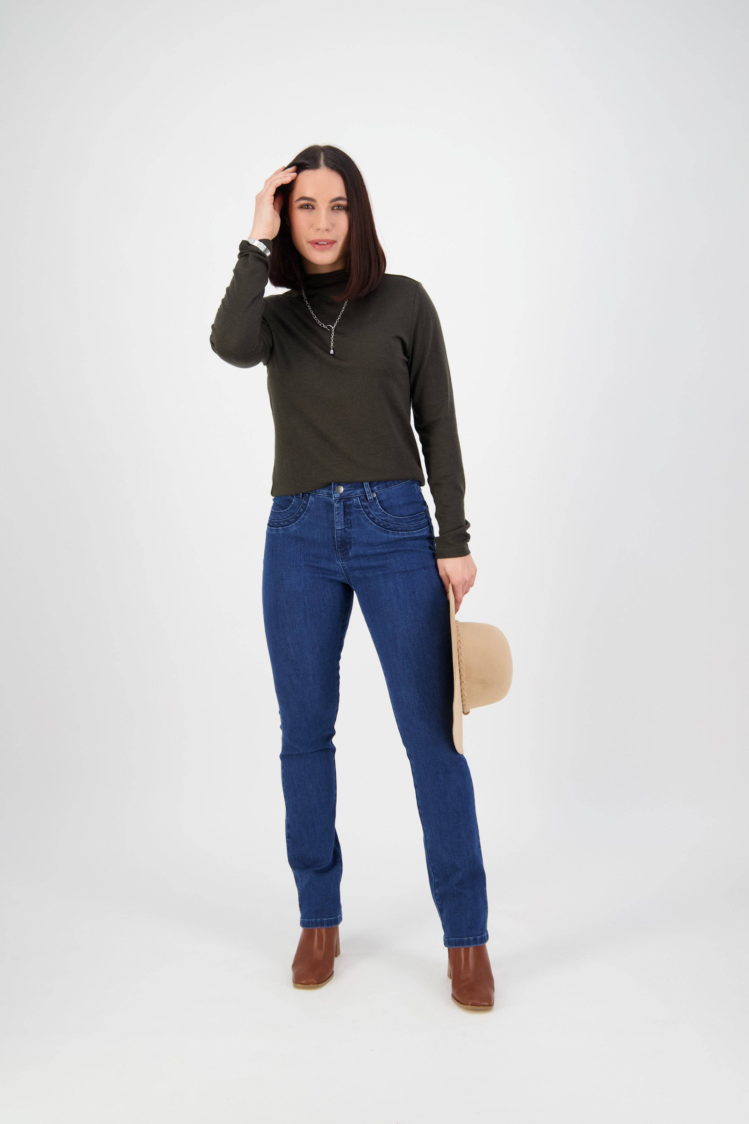 A woman stands against a white background, wearing a dark green long-sleeve top, Vassalli Studio Jean in blue denim, and brown boots. She holds a beige hat in one hand and touches her hair with the other while looking at the camera.