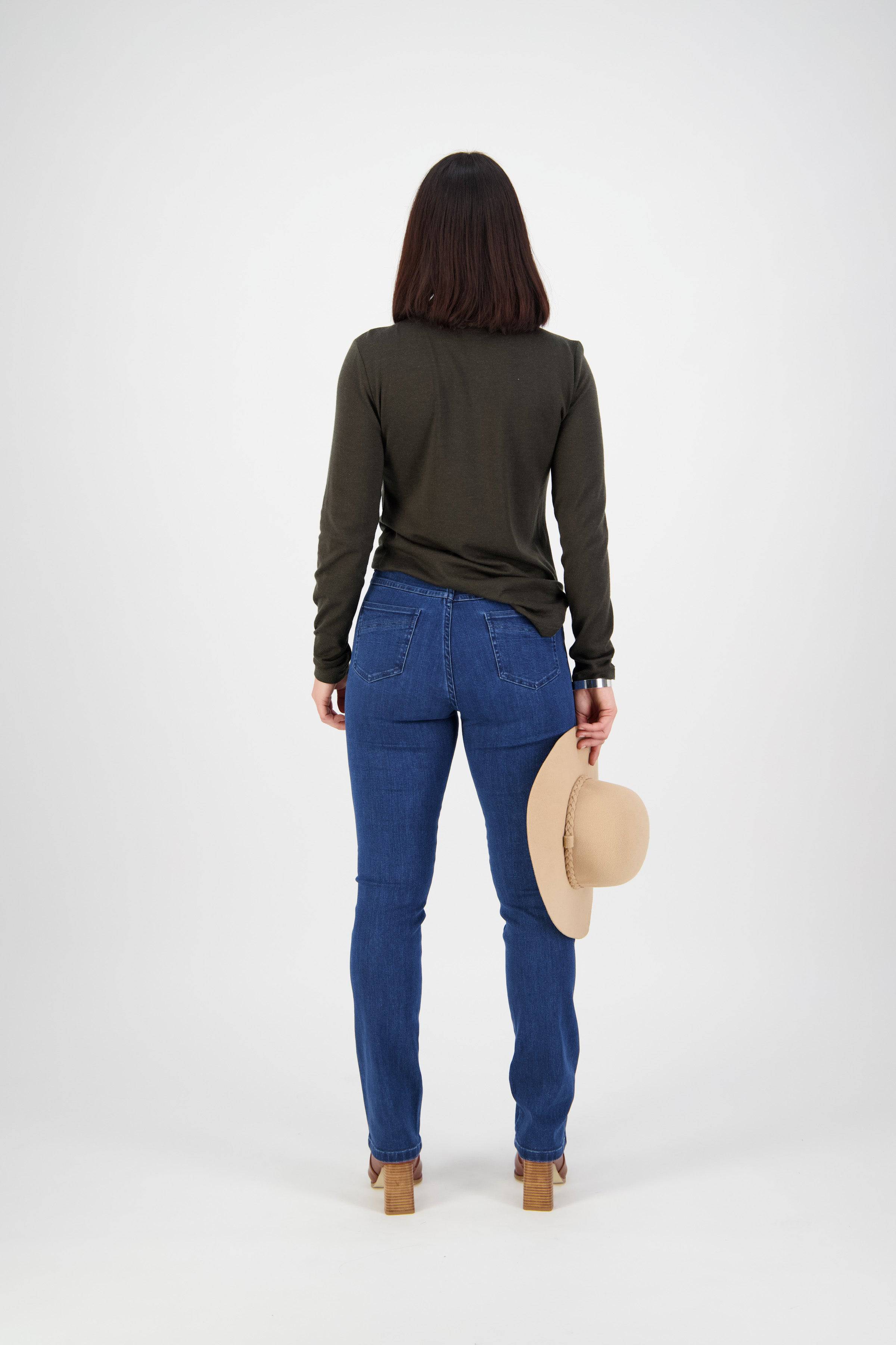 A woman faces away against a plain white background, wearing a dark long-sleeve shirt, Vassalli Studio Jean - Blue Denim, heeled shoes, and holding a beige hat in her right hand.