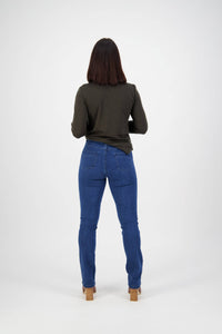 A woman with shoulder-length brown hair faces away, wearing a long-sleeve dark green top, Vassalli Studio Jean in blue denim, and heeled sandals against a plain white background.