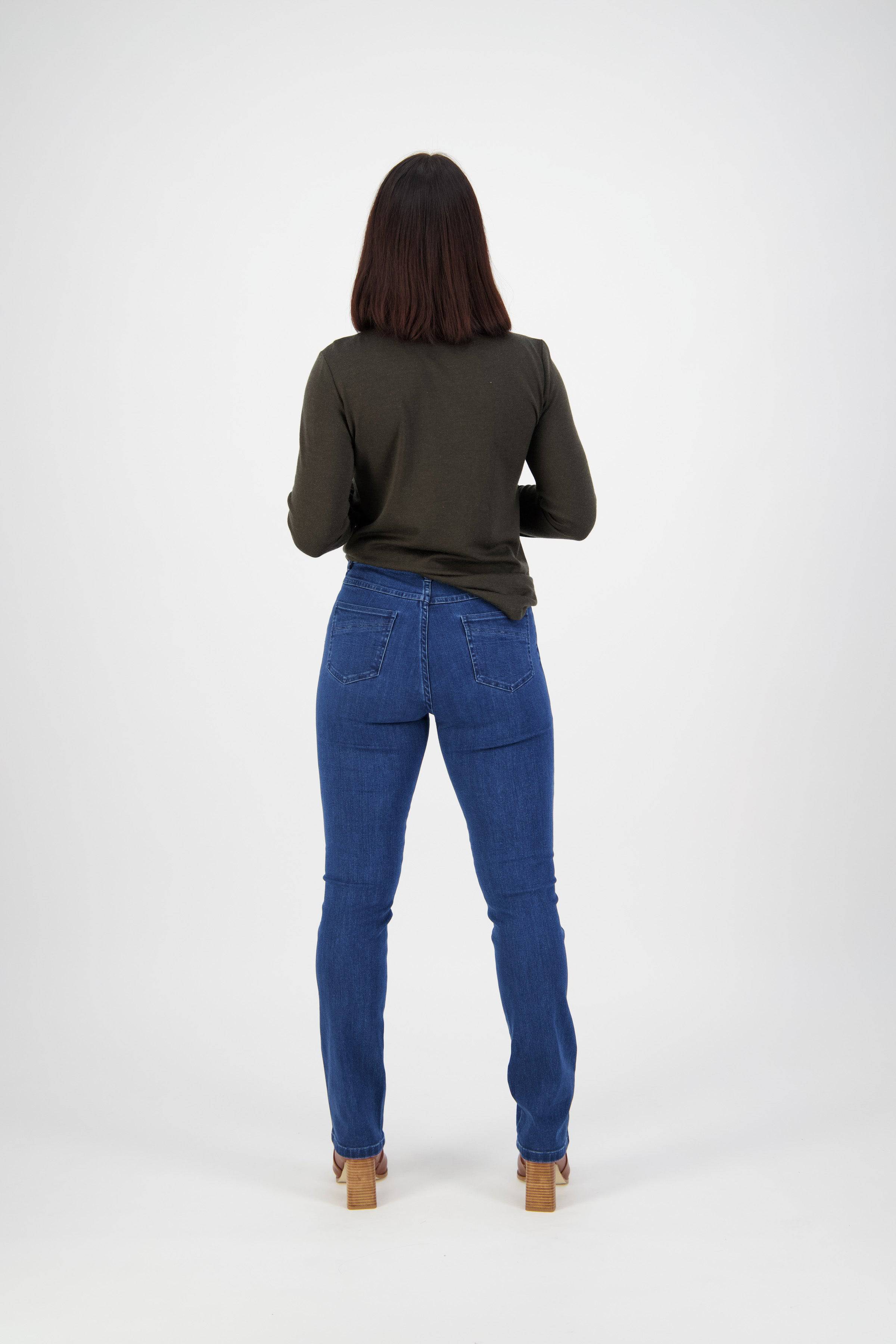 A woman with shoulder-length brown hair faces away, wearing a long-sleeve dark green top, Vassalli Studio Jean in blue denim, and heeled sandals against a plain white background.