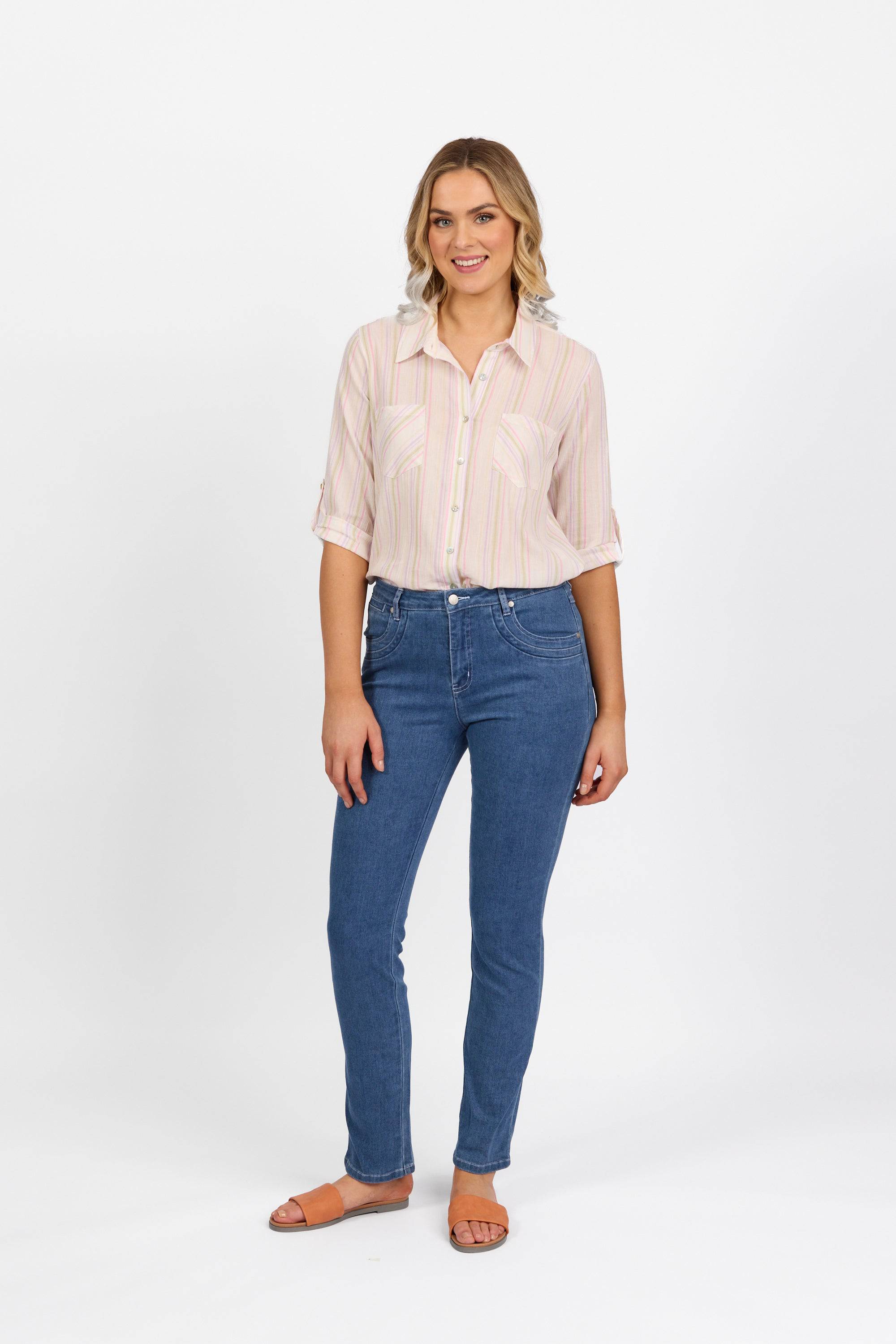 A woman smiles against a white background, wearing Vassalli's Studio Jean in Blue Hue Denim, a light-striped button-up shirt, and tan sandals. Her hair is styled in loose waves.