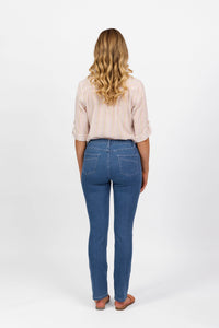 A woman with long wavy blonde hair, wearing a light pink striped shirt and Vassalli Studio Jean - Blue Hue Denim, stands in sandals facing away on a plain white background.