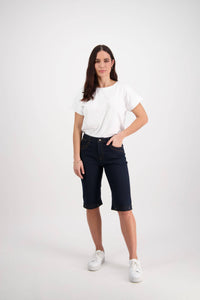 A woman stands against a white background, wearing Vassalli's Custom Short - Indigo Denim, a stretch-fabric straight-leg short, paired with a white short-sleeve t-shirt and white sneakers. One hand is in her pocket as she looks straight ahead.