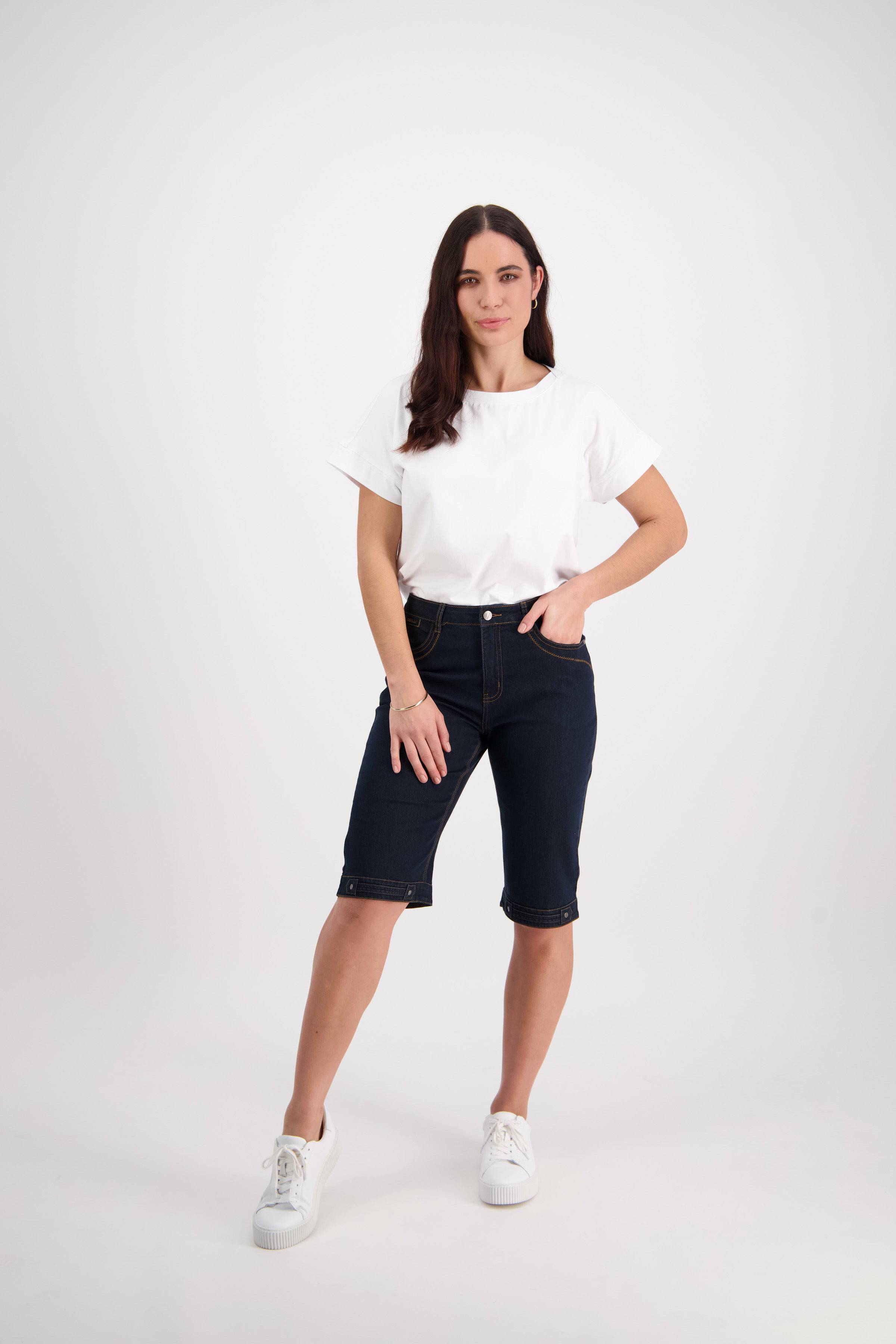 A woman with long brown hair stands against a white background, wearing a plain white t-shirt, Vassalli's Custom Short - Indigo Denim made from stretch fabric, and white sneakers. She poses with one hand in her pocket and a neutral expression.