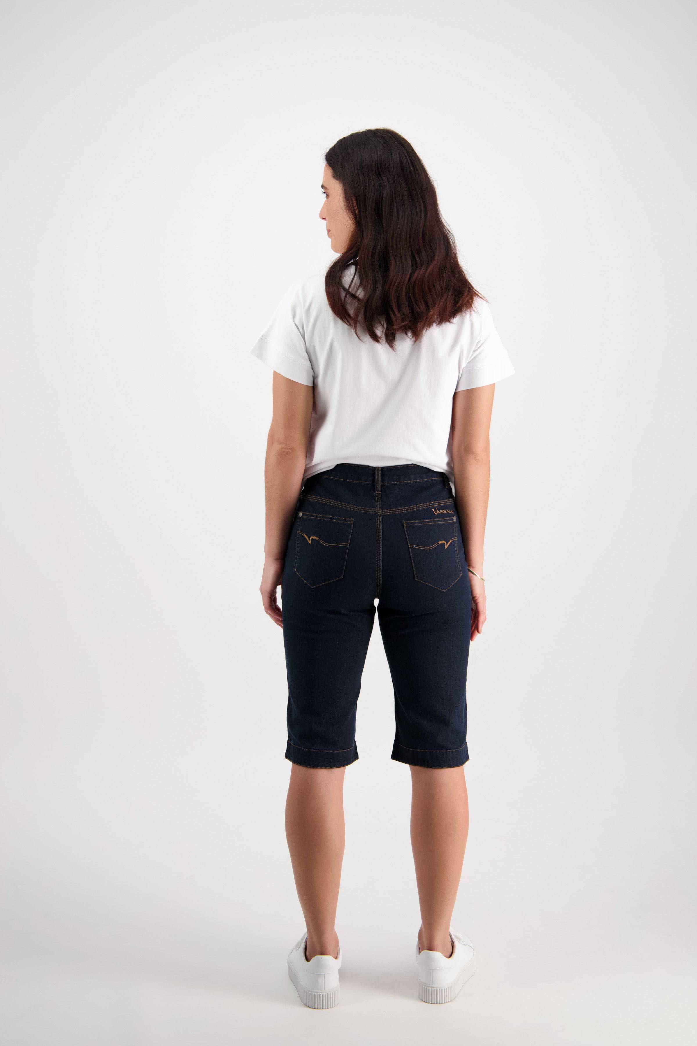 A woman with wavy brown hair, wearing a white t-shirt and Vassalli Custom Short - Indigo Denim, paired with white sneakers, stands facing away from the camera against a plain white background.