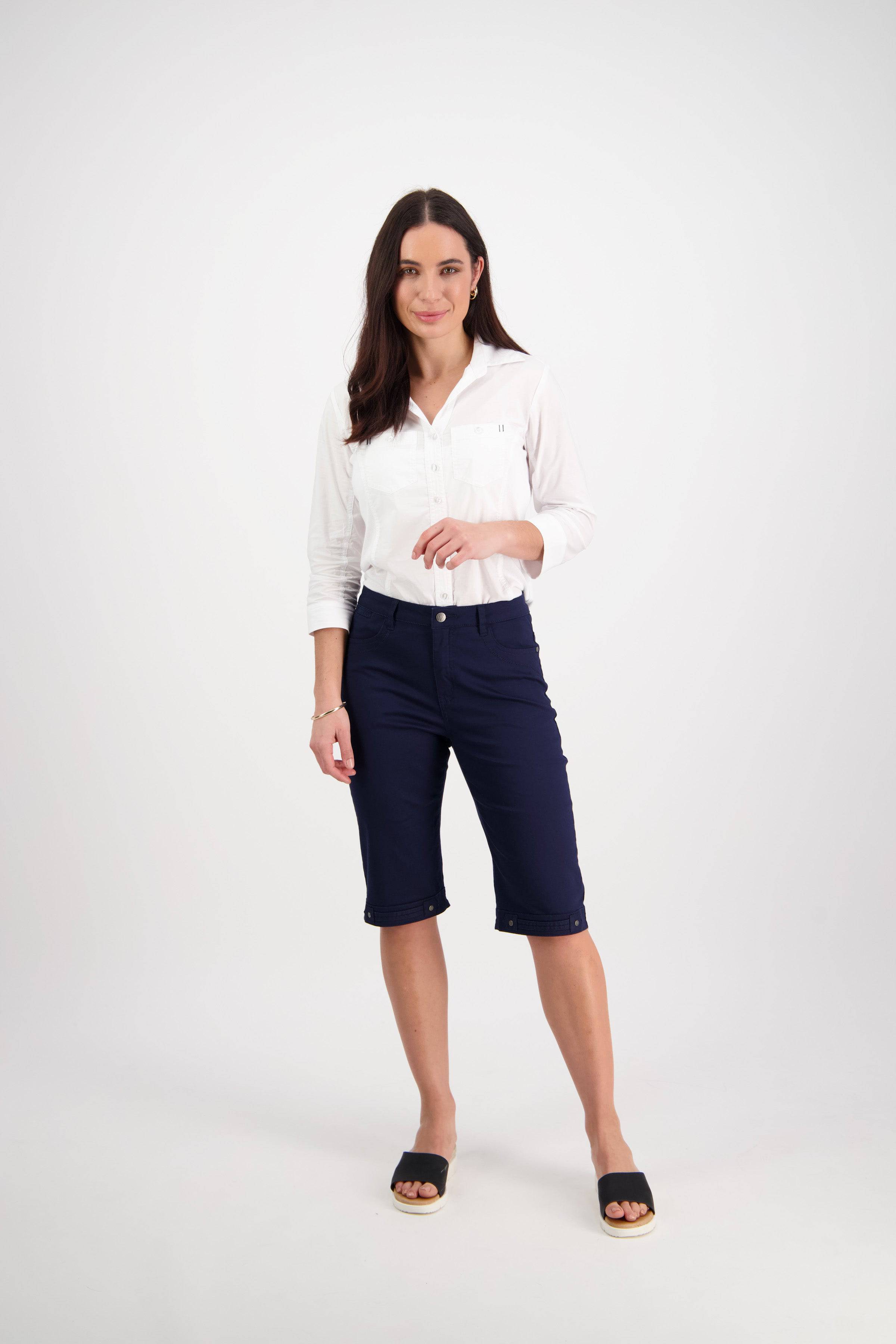 A woman with long dark hair stands against a white background, wearing a white button-up shirt, Vassalli Custom Lightweight Short in navy, and black slip-on sandals—a comfortable summer look. She faces forward with a slight smile.