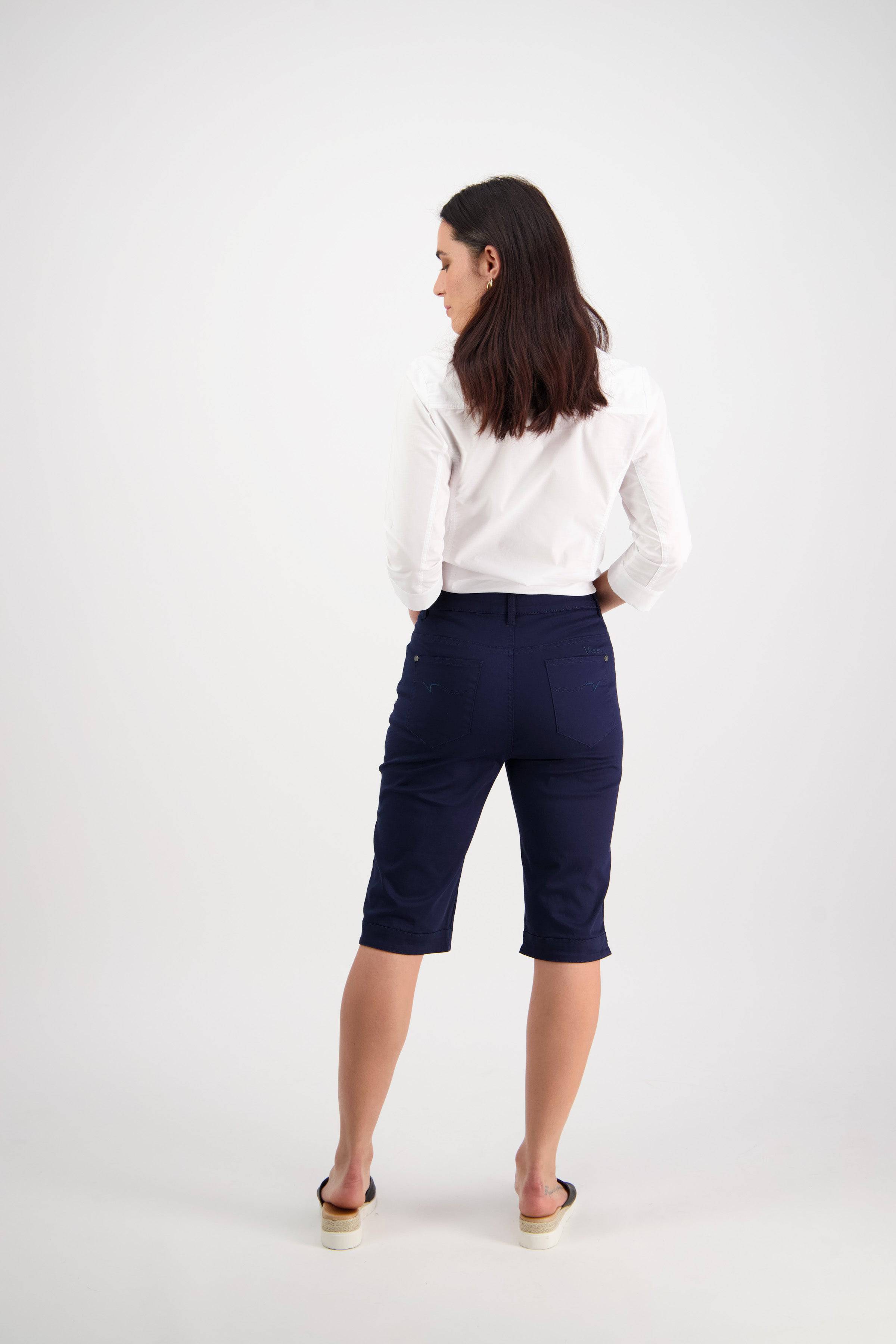 A woman with dark hair, wearing a white long-sleeve shirt, Vassalli Custom Lightweight Short in navy, and sandals stands facing away from the camera against a plain white background.