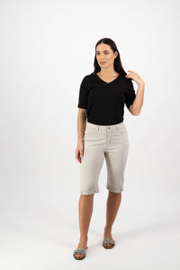 A woman with long dark hair stands against a white background wearing Vassalli’s Custom Lightweight Short in Pumice and a black V-neck top, touching her hair and showcasing a relaxed summer look.