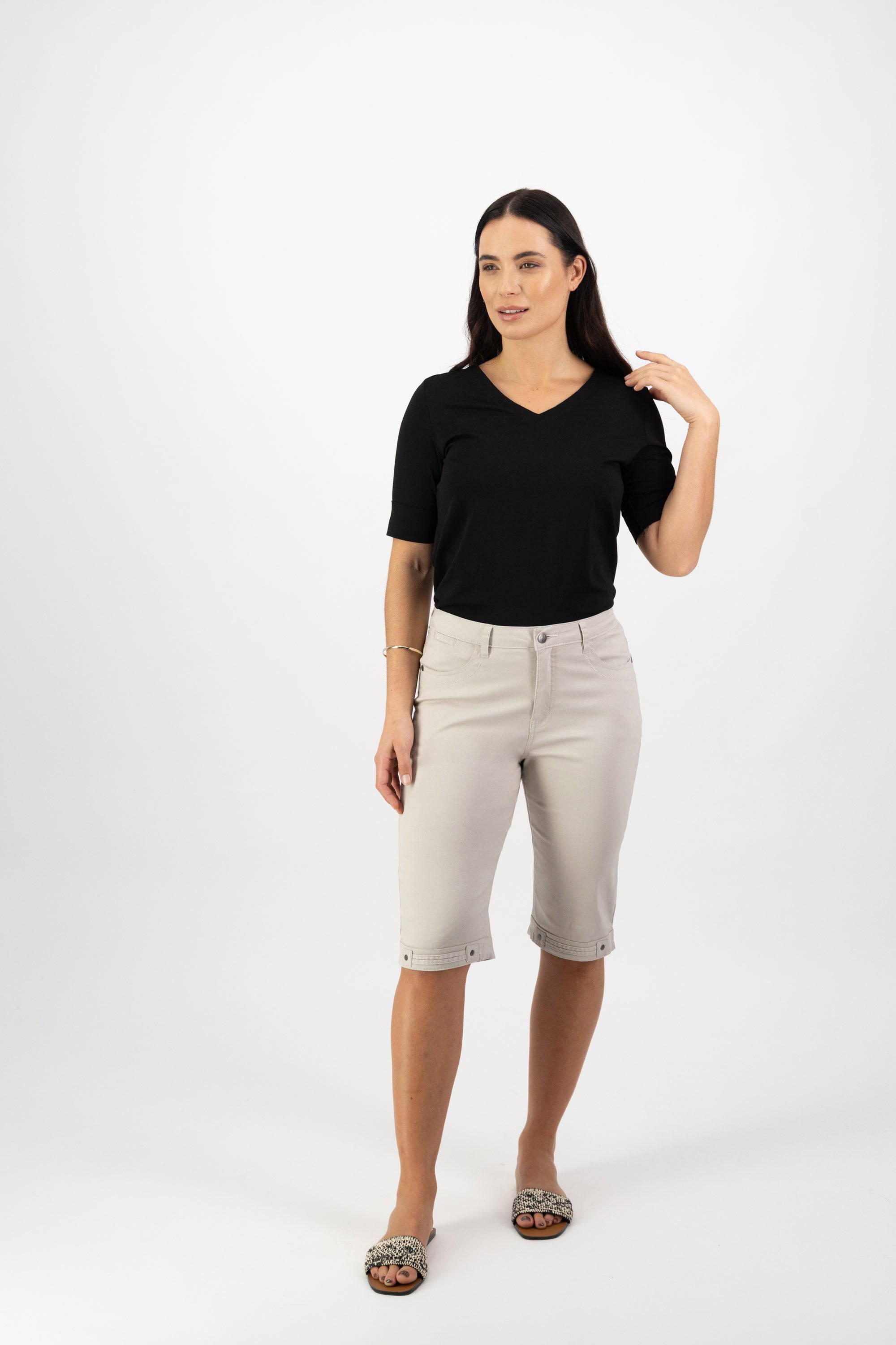 A woman with long dark hair stands against a white background wearing Vassalli’s Custom Lightweight Short in Pumice and a black V-neck top, touching her hair and showcasing a relaxed summer look.