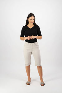 A woman with long dark hair stands against a white background, wearing a black top and Vassalli’s Custom Lightweight Short in Pumice—perfect for summer. Leopard-print slip-ons complete her look as she glances to the side, hands loosely clasped.