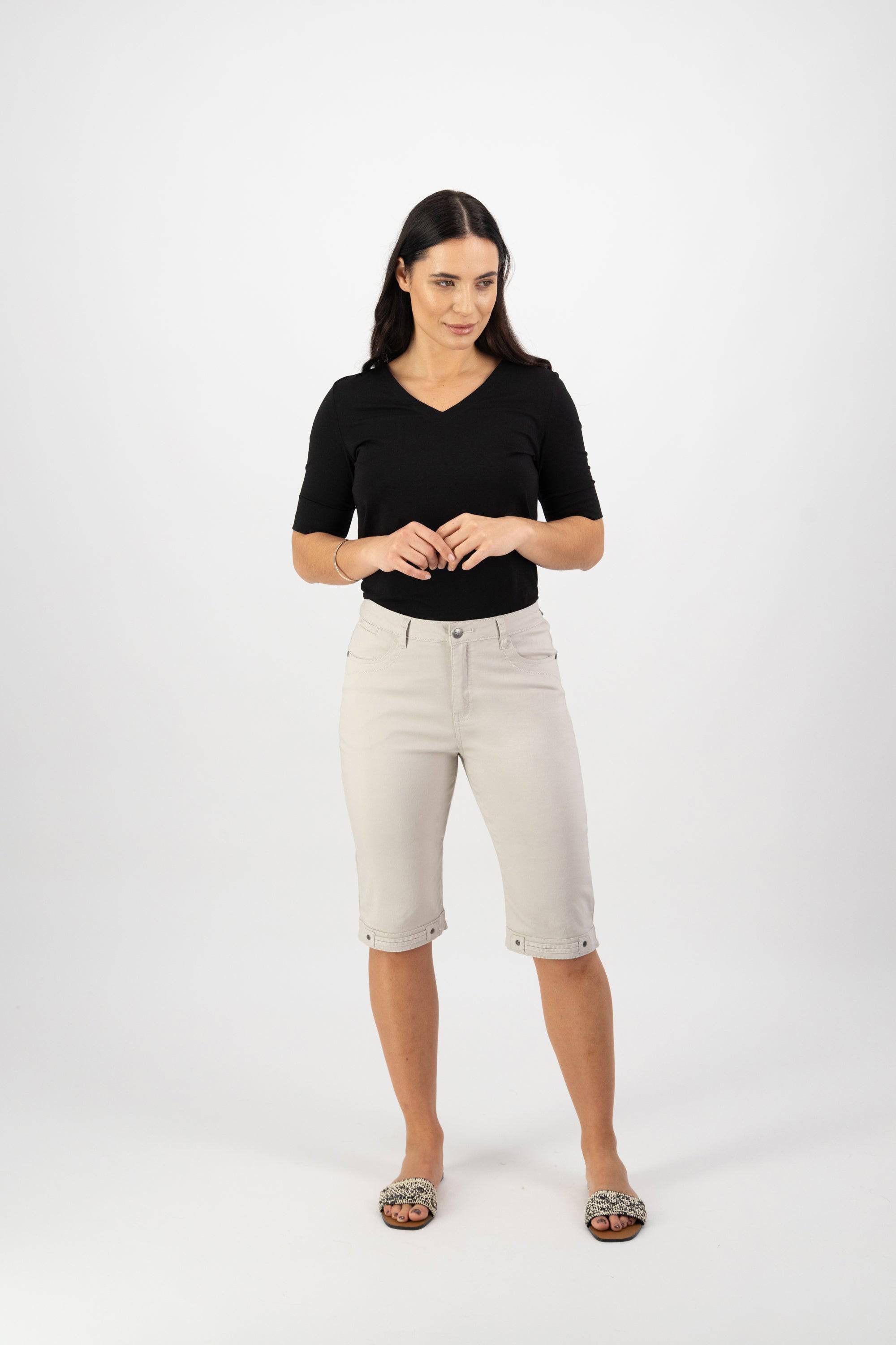A woman with long dark hair stands against a white background, wearing a black top and Vassalli’s Custom Lightweight Short in Pumice—perfect for summer. Leopard-print slip-ons complete her look as she glances to the side, hands loosely clasped.