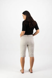 A woman with long brown hair stands facing away, wearing a black top, Vassalli Custom Lightweight Short in Pumice for a comfortable look, and flat shoes, her hands in her pockets against a plain white background—perfect for summer.