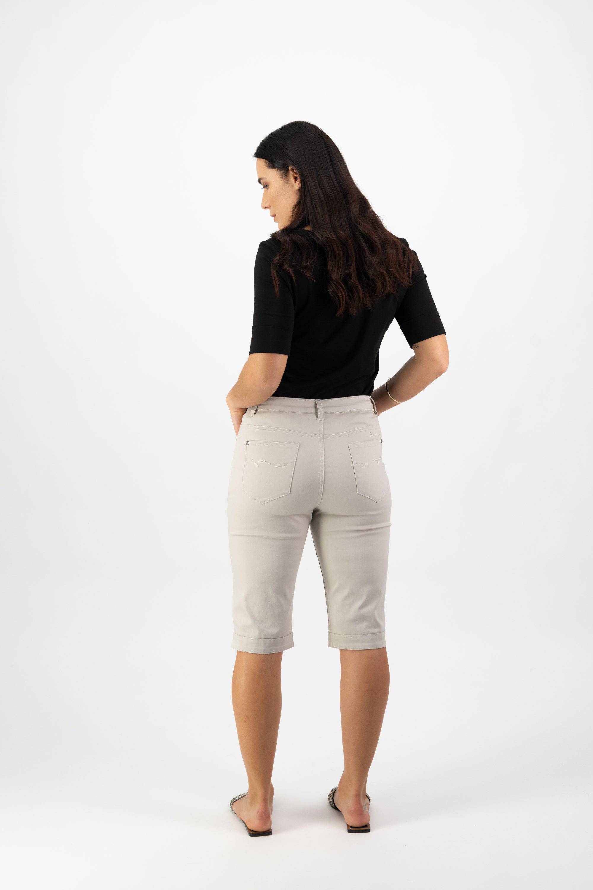 A woman with long brown hair stands facing away, wearing a black top, Vassalli Custom Lightweight Short in Pumice for a comfortable look, and flat shoes, her hands in her pockets against a plain white background—perfect for summer.