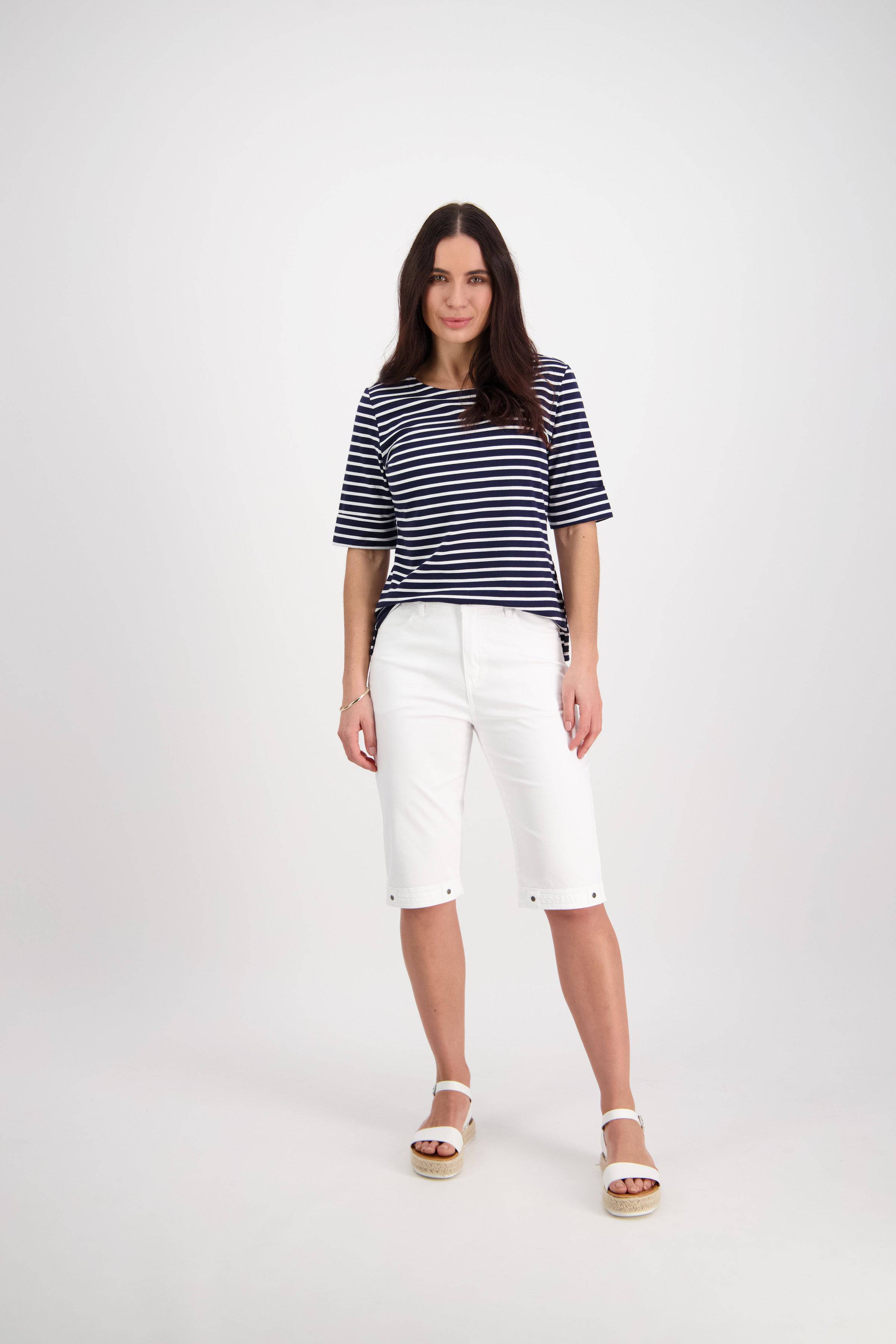 A woman stands in a studio wearing Vassalli’s Custom Lightweight Short in white, paired with a navy and white striped top and sandals. Her relaxed, confident look embodies comfortable summer style.