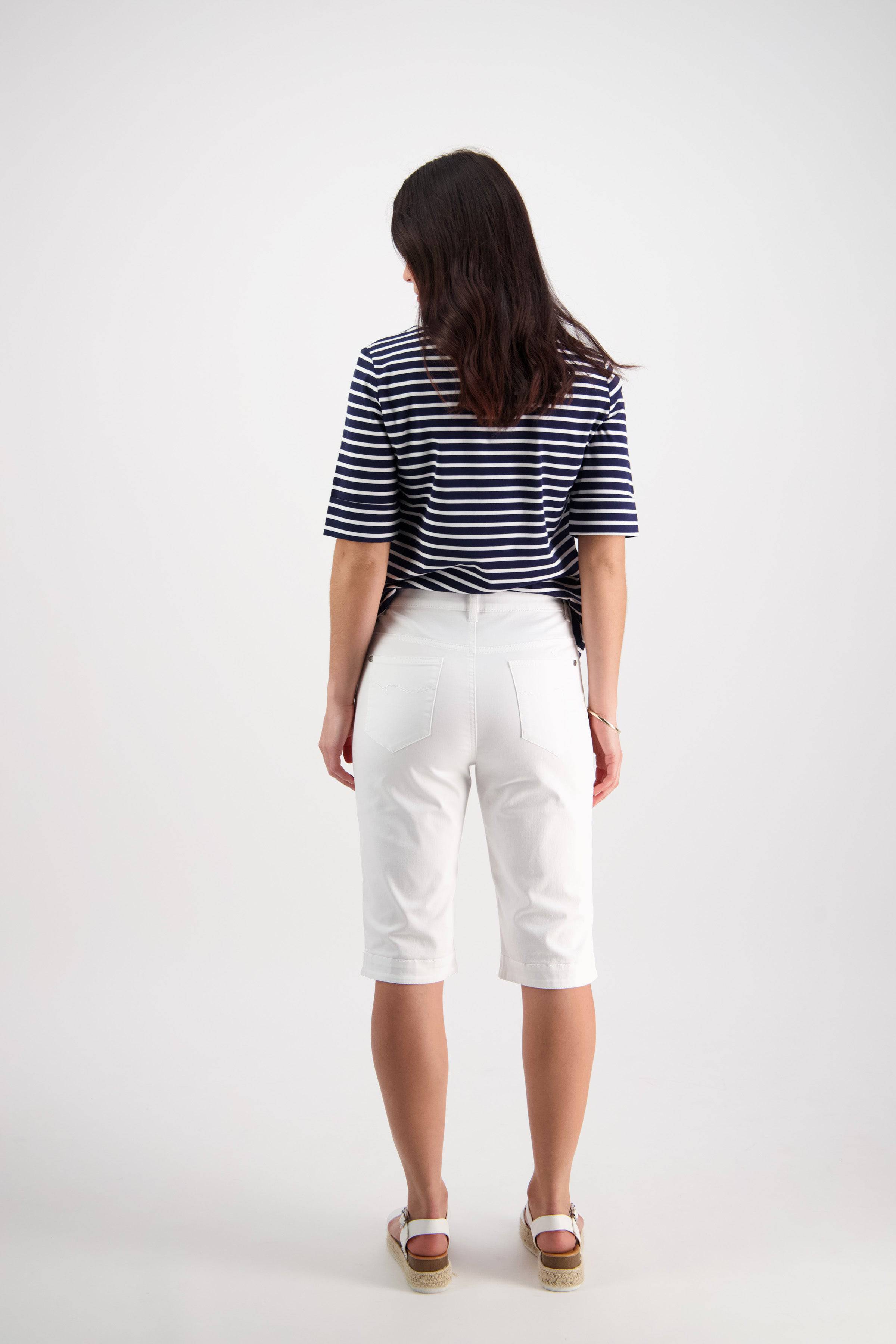 A woman stands facing away in a navy and white striped short-sleeve shirt, Vassalli Custom Lightweight Short in white for comfort, and wedge sandals against a plain white background.