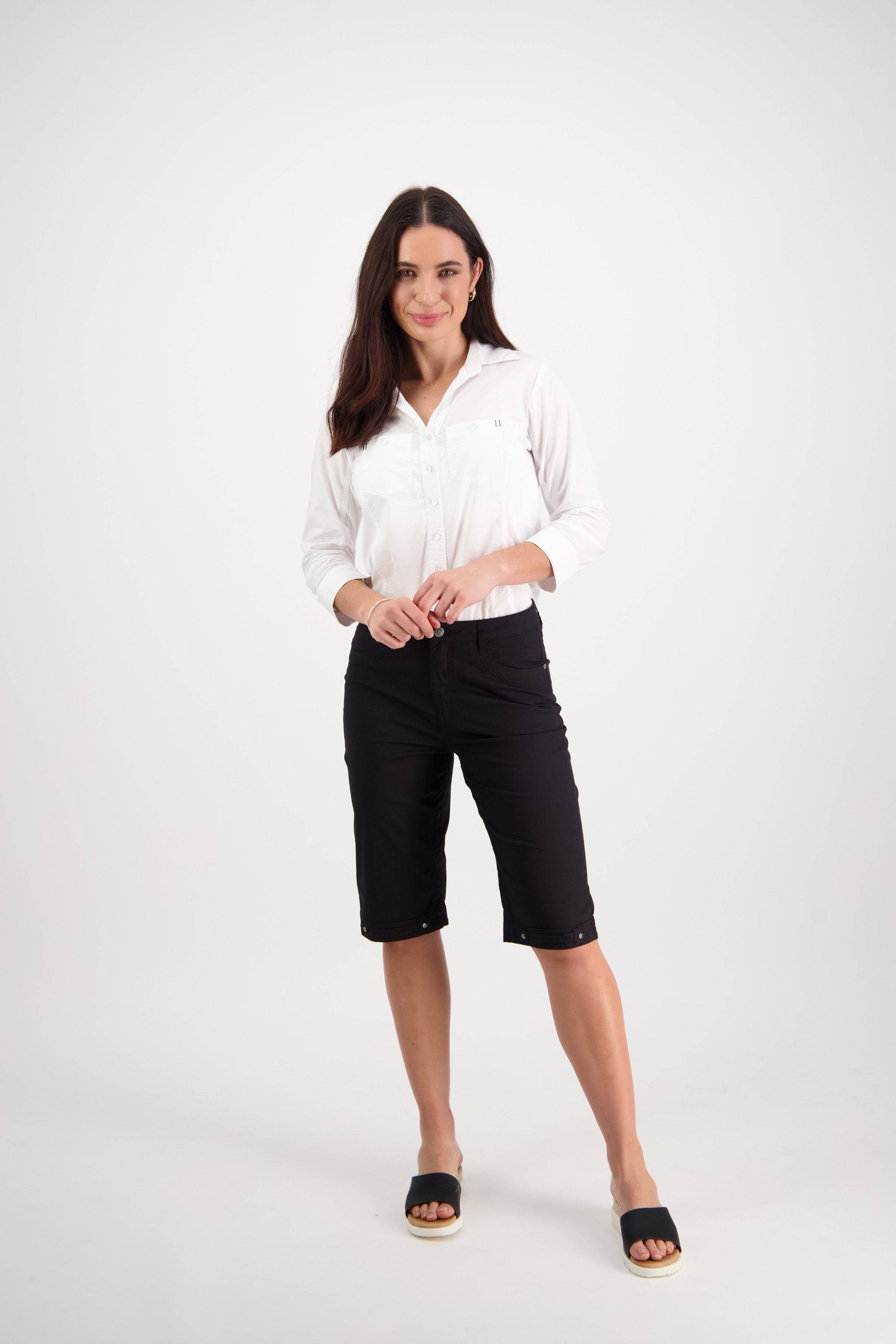 A woman with long brown hair stands against a plain white background, wearing the Vassalli Custom Poplin Short in black, a white button-up shirt, and slide sandals. She looks at the camera with a slight smile.