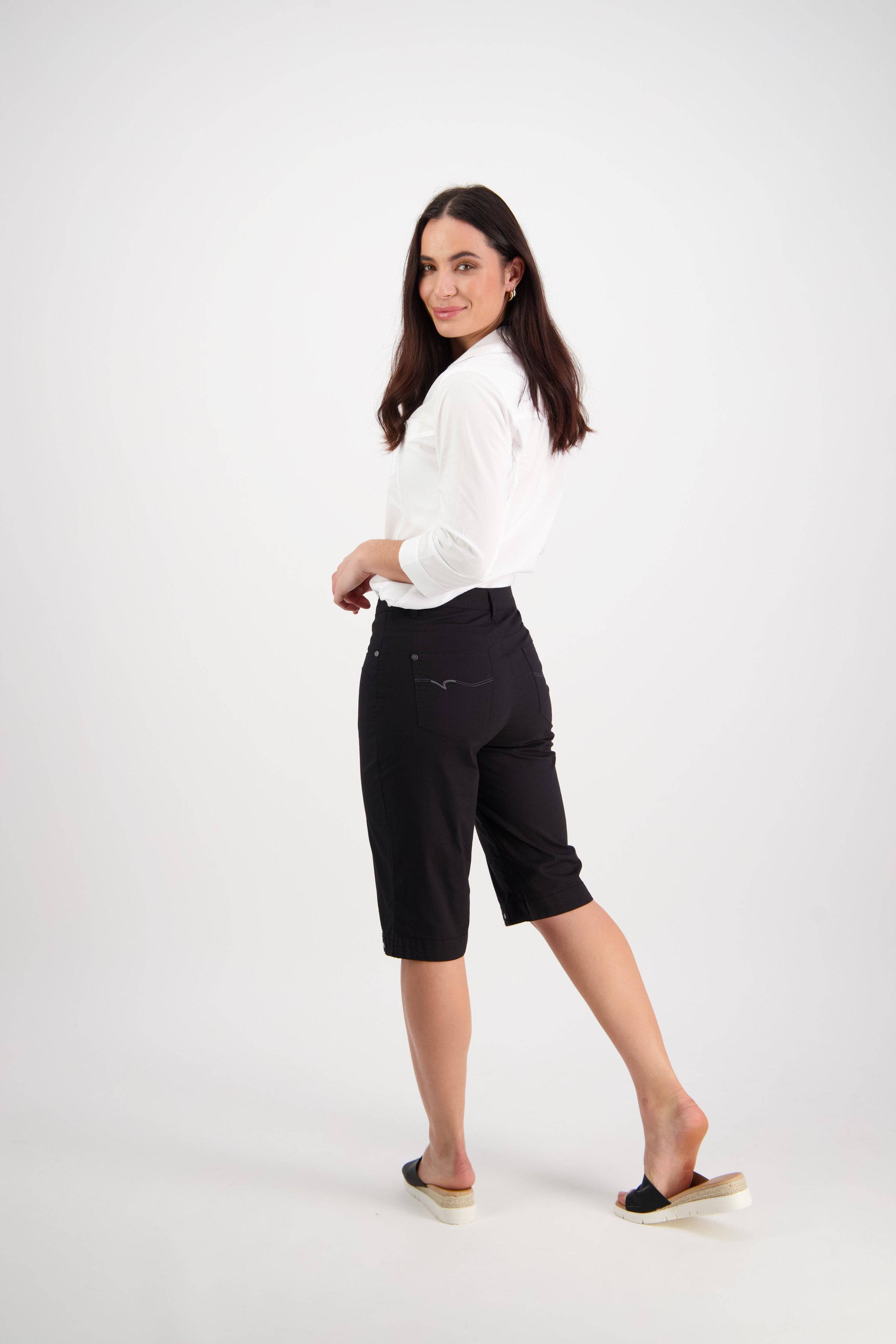 A woman with long dark hair, wearing a white long-sleeve top and Vassalli Custom Poplin Short in black, stands on one leg and smiles over her shoulder in a bright studio setting.