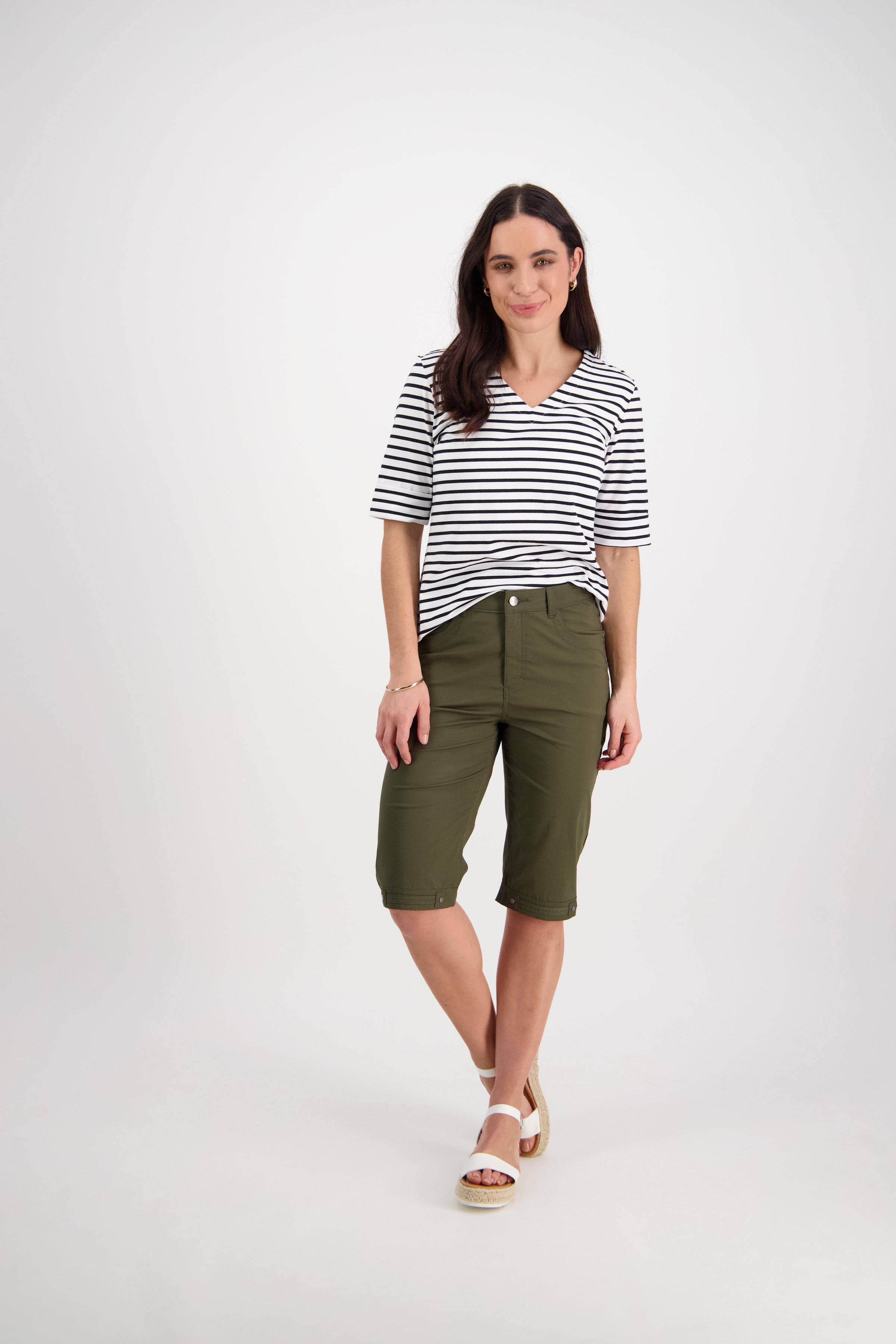 A woman with long dark hair stands against a white background wearing a black-and-white striped short-sleeve shirt, Vassalli Custom Poplin Short in khaki, and white sandals for a relaxed, versatile look.