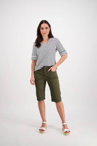 A woman with long dark hair poses confidently against a white background, wearing a black-and-white striped shirt, Vassalli Custom Poplin Short in khaki, and white sandals. She stands relaxed with one hand in her pocket.