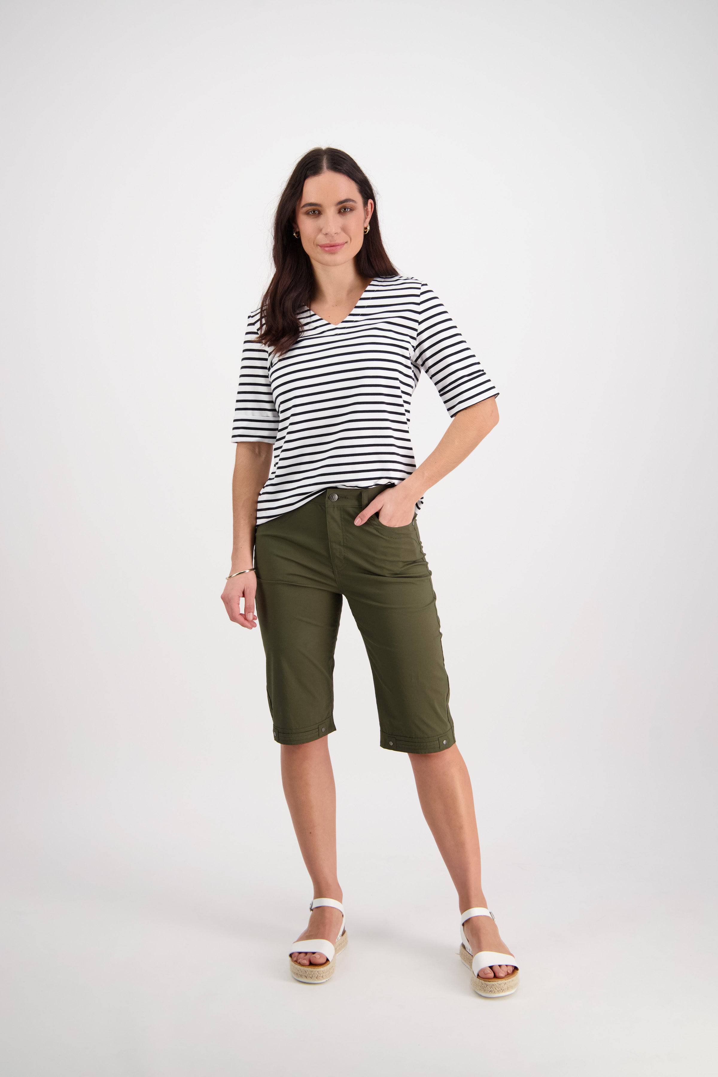 A woman with long dark hair poses confidently against a white background, wearing a black-and-white striped shirt, Vassalli Custom Poplin Short in khaki, and white sandals. She stands relaxed with one hand in her pocket.