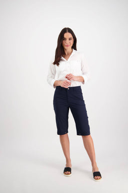 A woman with long dark hair smiles slightly as she poses against a white background, wearing Vassalli's Custom Poplin Short in Navy paired with a white long-sleeve blouse and black slide sandals.