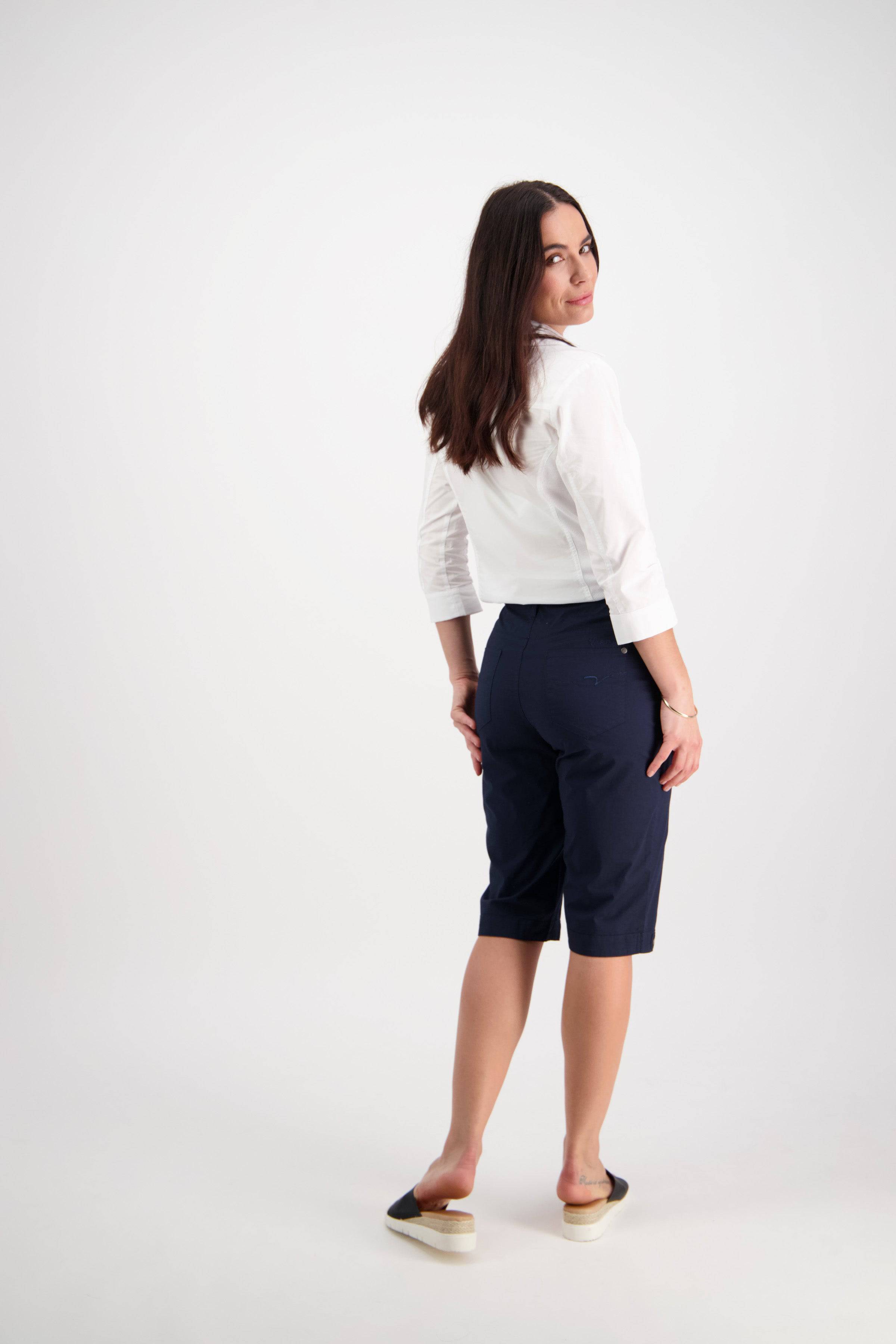 A woman with long brown hair, facing away and looking at the camera, wears a white shirt, black flats, and Vassalli's Custom Poplin Short in Navy—a tailored short with cuffed hems—set against a plain white background.