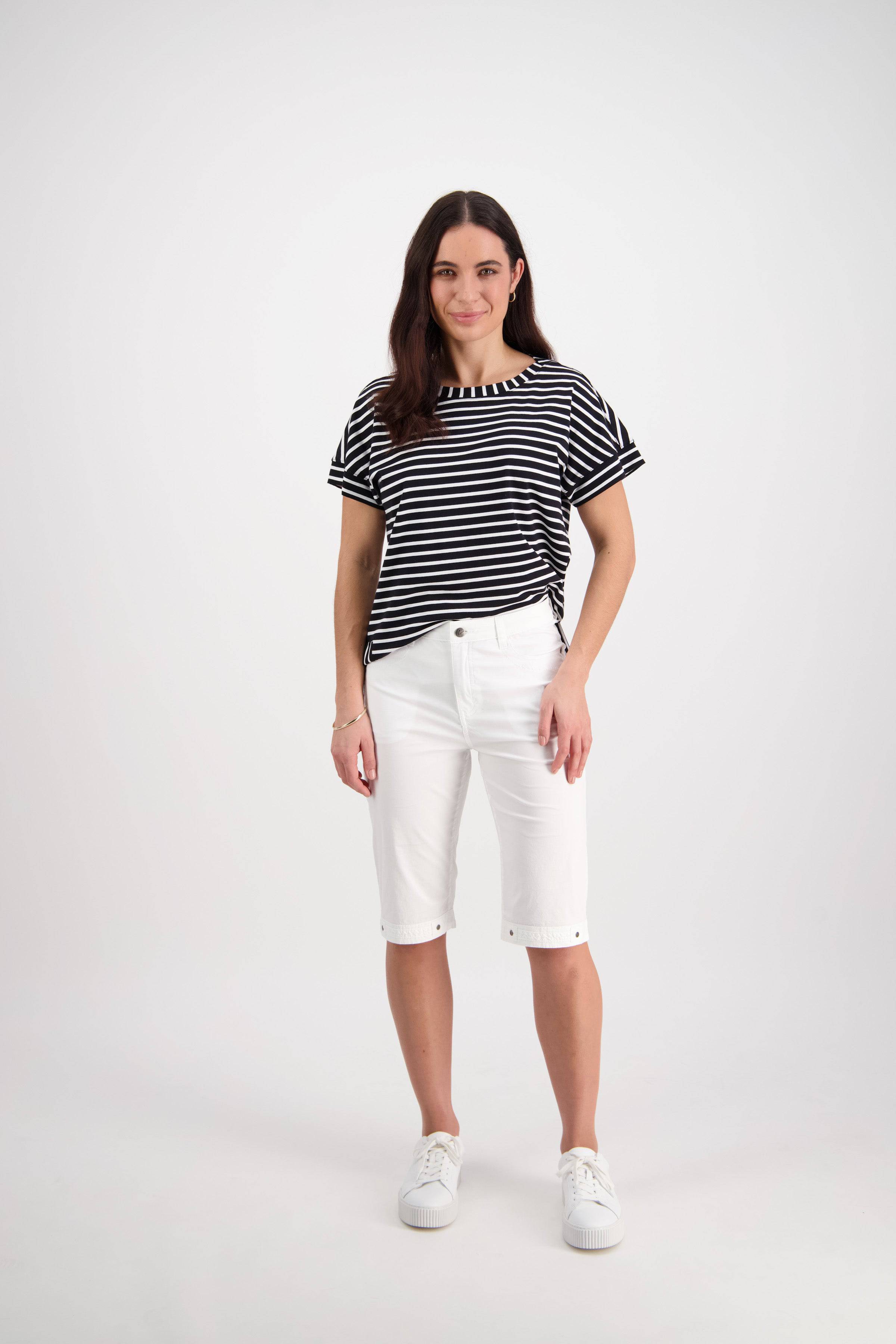 A woman with long brown hair poses casually against a white background, wearing Vassalli's Custom Poplin Short in white, a black and white striped t-shirt, and white sneakers.