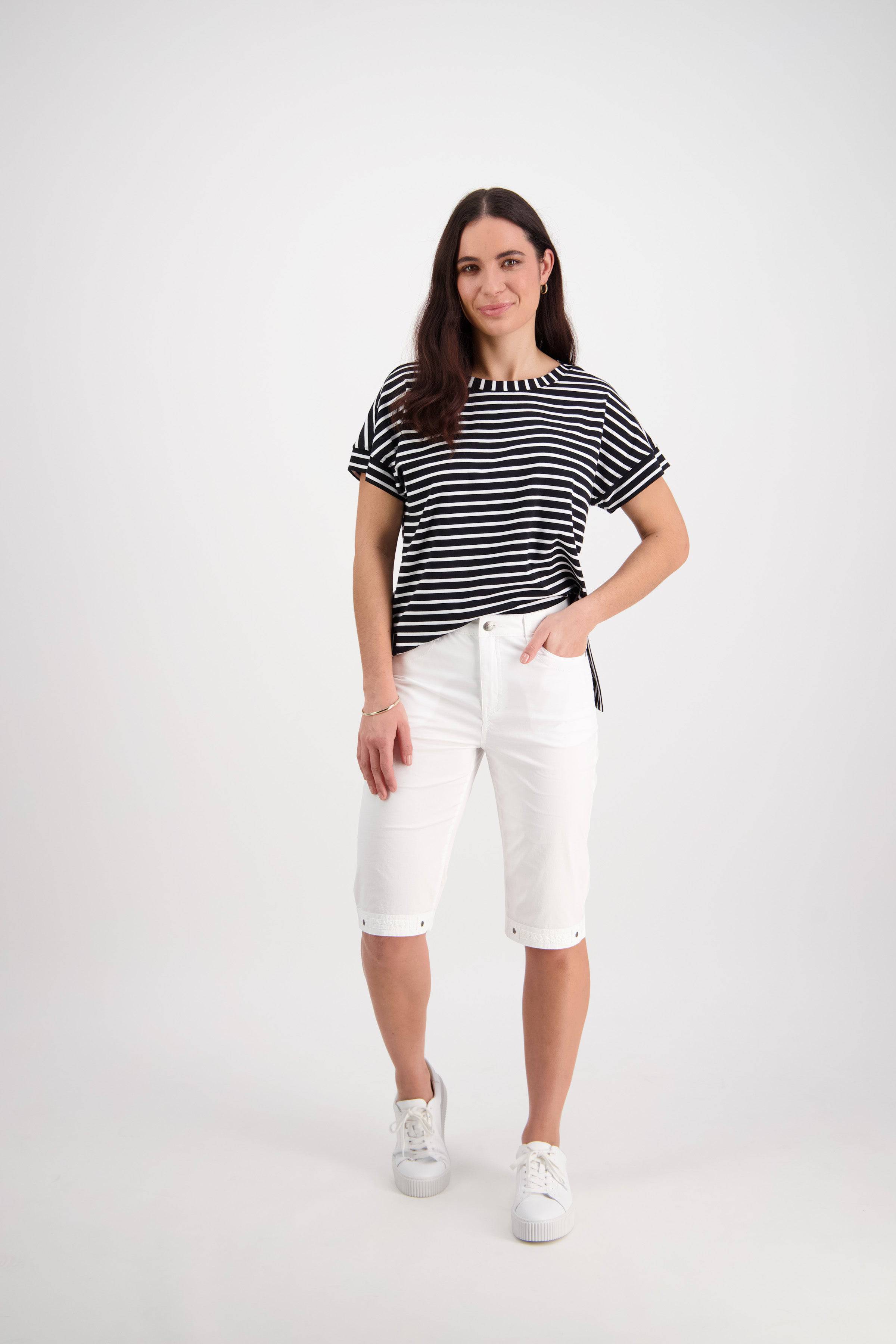 A woman with long brown hair smiles with one hand in her pocket, wearing Vassalli’s Custom Poplin Short in White, a black and white striped short-sleeve top, and white sneakers against a plain white background.