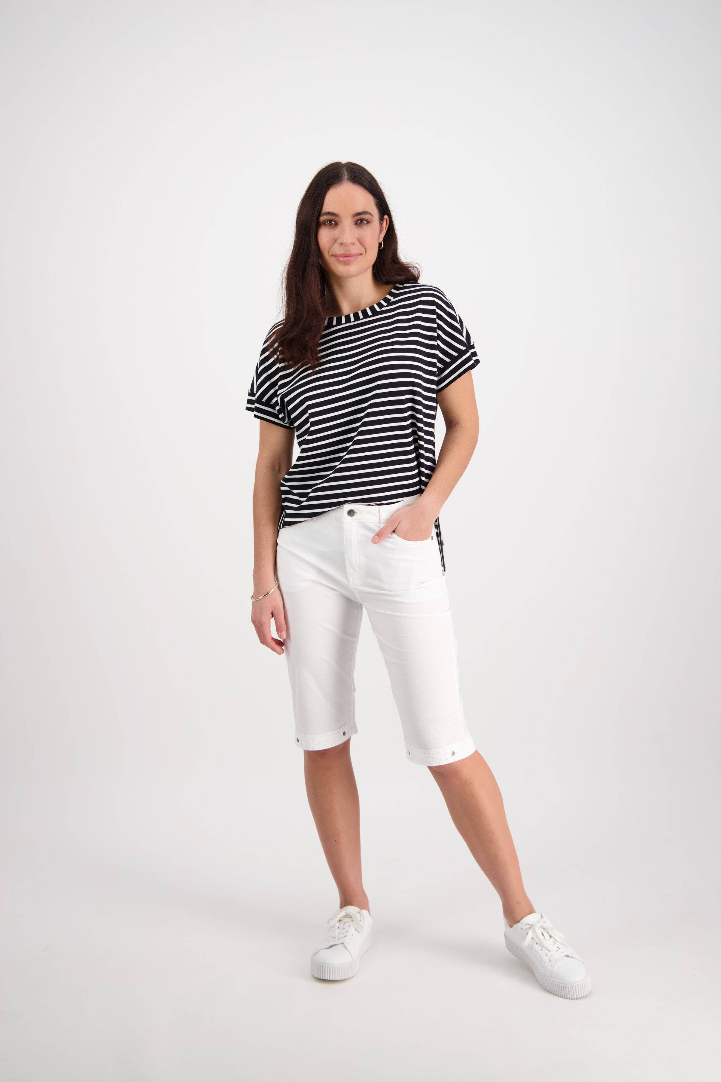 A woman stands against a white background, wearing a black and white striped t-shirt, Vassalli Custom Poplin Short in white, and white sneakers. She has one hand in her pocket and is smiling slightly.