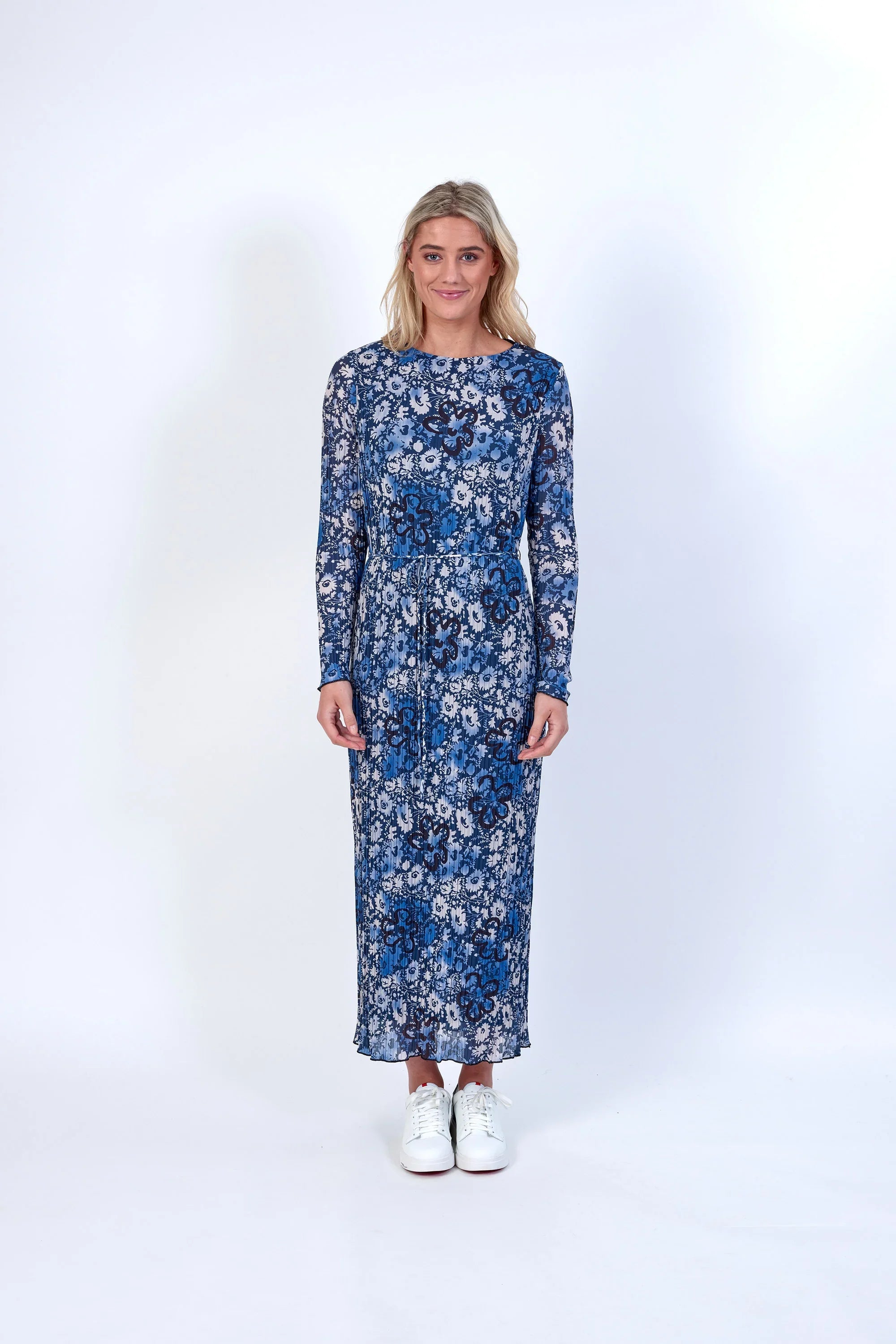 Woman in a blue floral long-sleeve midi dress with white sneakers standing against a white background