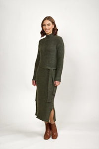 Woman in olive green knit long-sleeve belted dress and brown ankle boots on white background