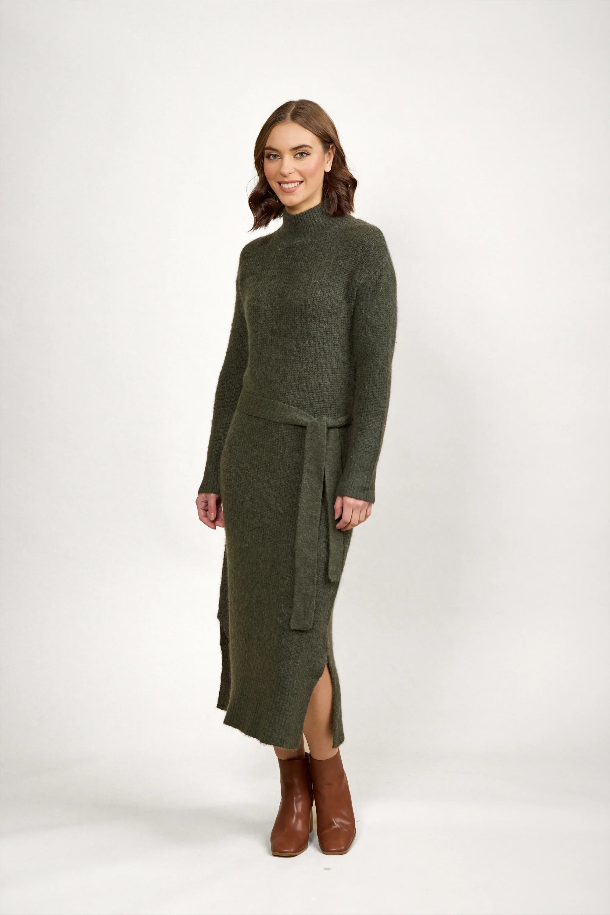 Woman in olive green knit long-sleeve belted dress and brown ankle boots on white background
