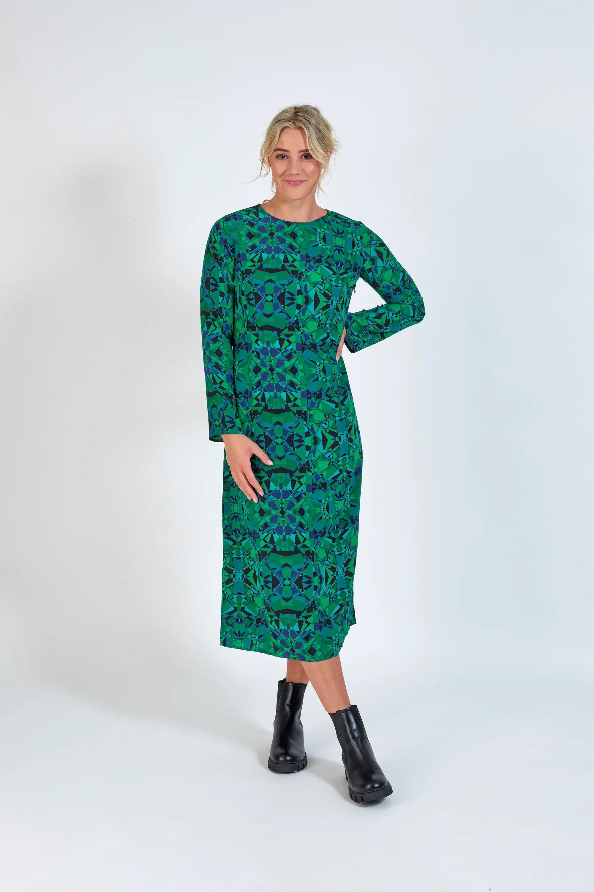 Woman wearing a green geometric long-sleeve midi dress with black ankle boots on a white background.