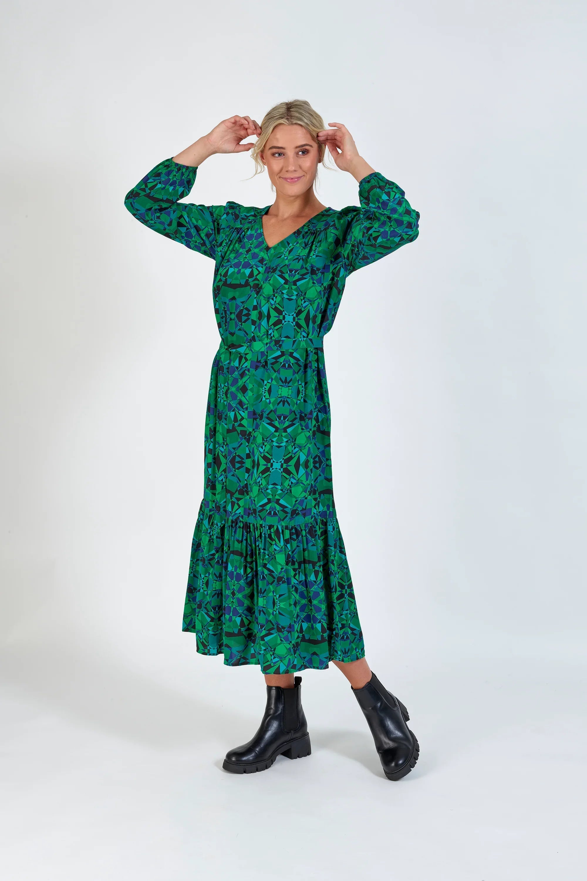 Woman in a green geometric print midi dress with long sleeves and black boots, studio background.