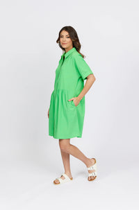 Woman wearing a bright green cocoon dress with short sleeves and pockets, styled with white slide sandals, on a plain background.