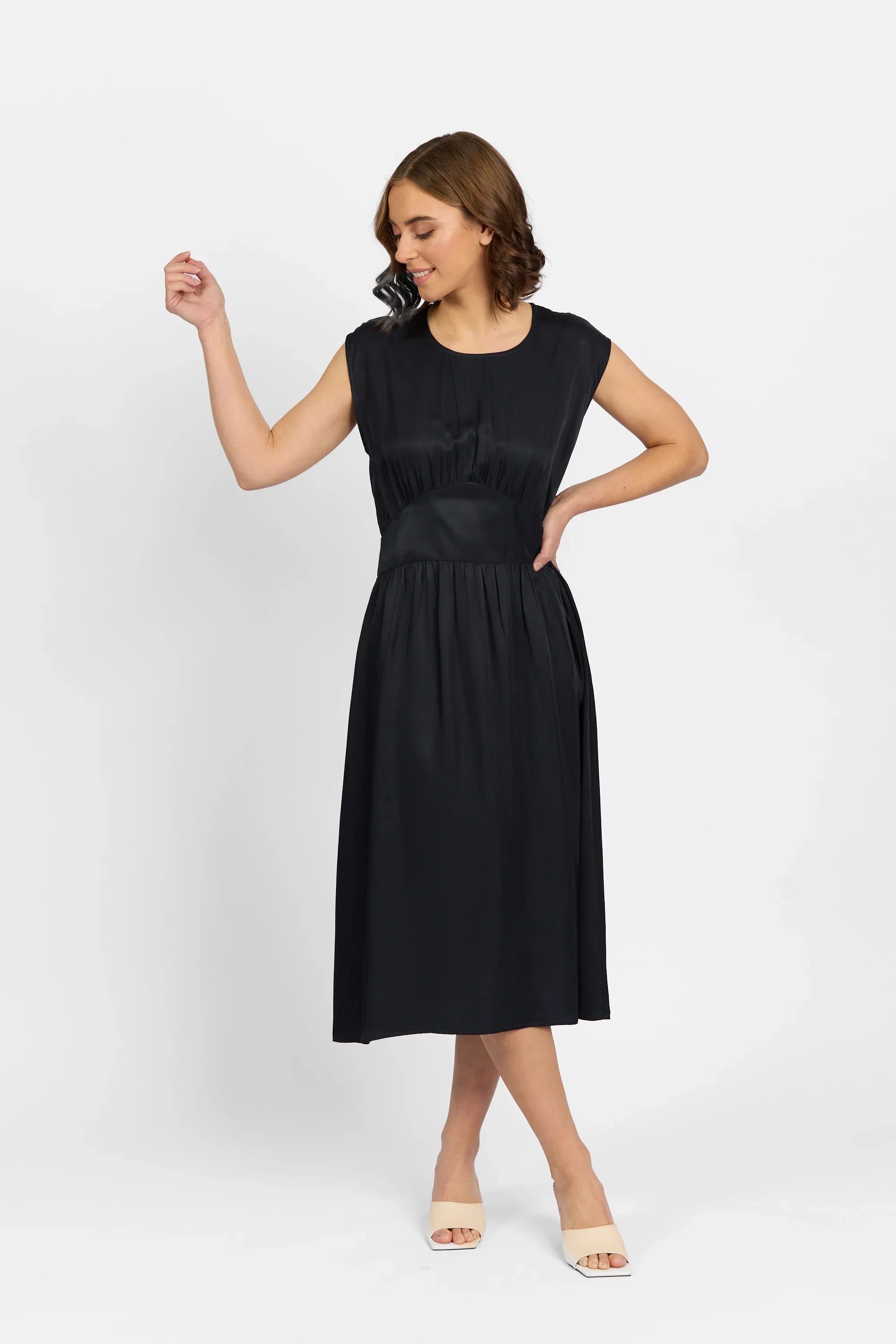 Woman wearing a sleeveless black midi dress with ruched waist, posing against a white background